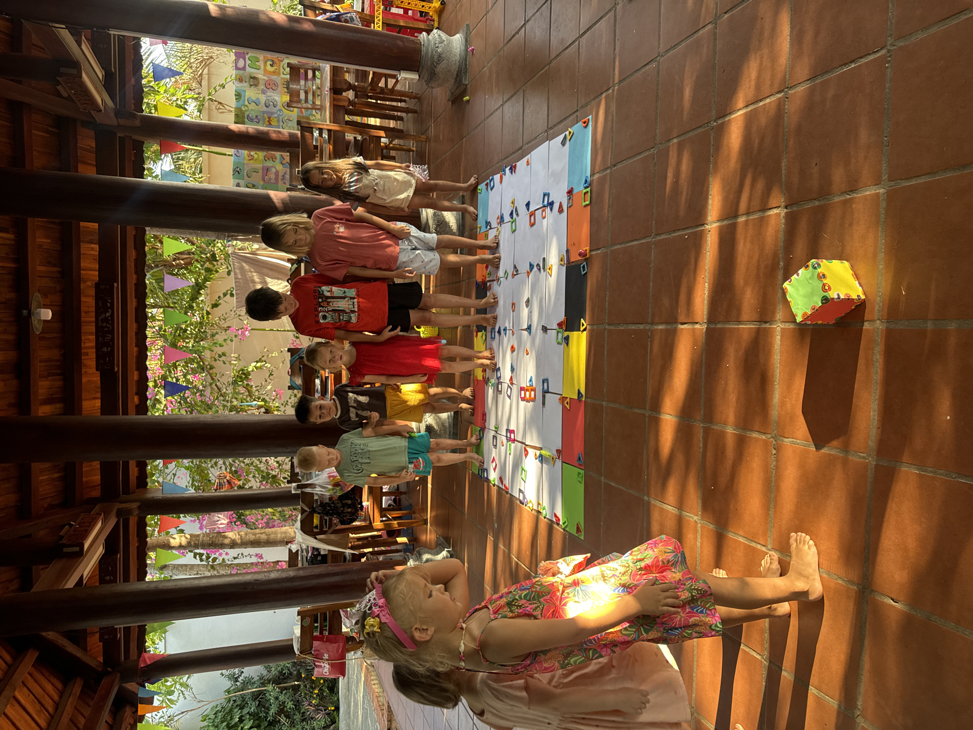 The image shows a group of children participating in an outdoor or semi-outdoor activity in Mũi Né, Vietnam. In the foreground, a young girl in a floral dress and tiara sits on the tiled floor, observing the scene. Behind her, a line of seven children of varying ages stands barefoot on a terracotta-tiled surface, facing a large, colorful mat or game board spread on the floor.
The mat is divided into a grid, adorned with numerous geometric shapes in various colors, including triangles, squares, and rectangles, suggesting an educational game. To the right of the mat, a large, soft, multi-colored cube, resembling a die or building block, rests on the floor. The children appear attentive and engaged, likely awaiting their turn or instructions for the game.
The setting is an open-air area, possibly a patio or covered walkway, characterized by robust wooden pillars and beams. Strong, angled shadows cast by the structure and subjects on the tiled floor indicate a bright, sunny day, likely morning or late afternoon. Lush green foliage and vibrant pink bougainvillea flowers are visible in the background, along with decorative flags or buntings, enhancing the festive and tropical atmosphere.
Visible text includes the word "EXPLORE" and graphic elements on a boy's red t-shirt, and "PLAYTIME" (partially obscured) on another boy's light green t-shirt. A partial brand name, "O'BON...ie," can be seen on a red shopping bag in the background. The overall scene depicts a lively and structured group activity in a warm, outdoor environment.