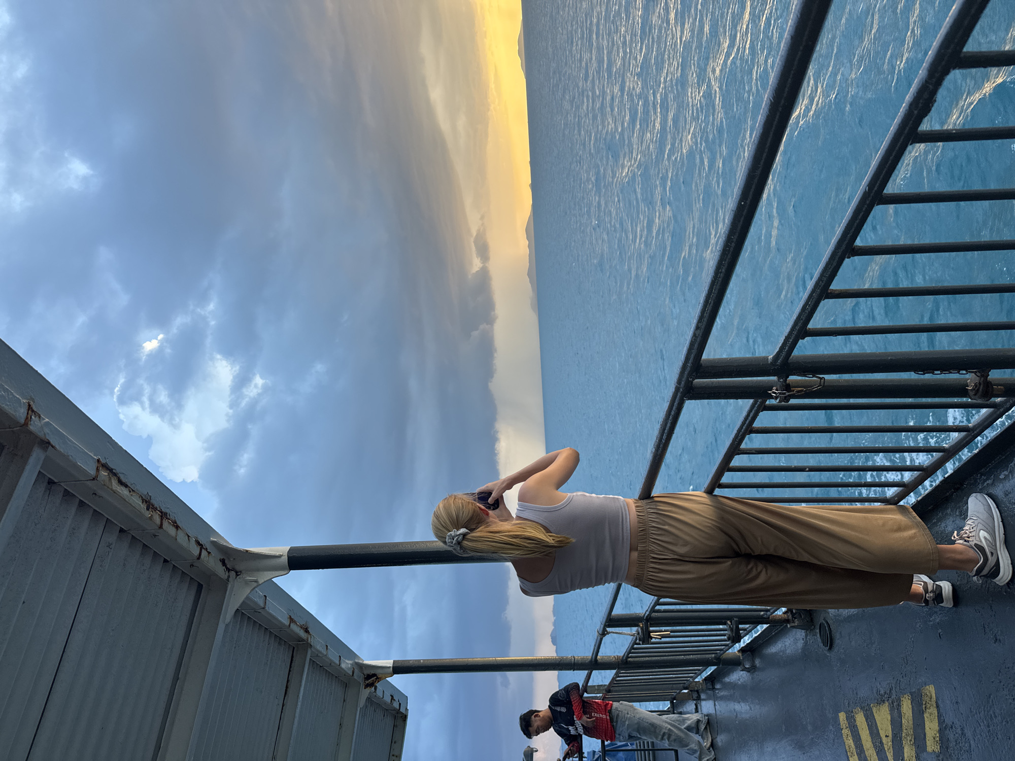 A woman with blonde hair pulled into a ponytail, wearing a white cropped tank top and tan wide-leg pants, stands on the deck of a ferry and takes a photo with her phone. She is facing away from the camera and looking out at the ocean. To her left, a man in a dark long-sleeved shirt with red and white accents and light-colored pants leans against the railing. The ferry deck is wet, with yellow markings on the floor. The ocean is a deep blue, and in the distance, islands are visible under a dramatic sky with dark clouds and patches of sunlight. The scene appears to be in Thailand, possibly at sunset.