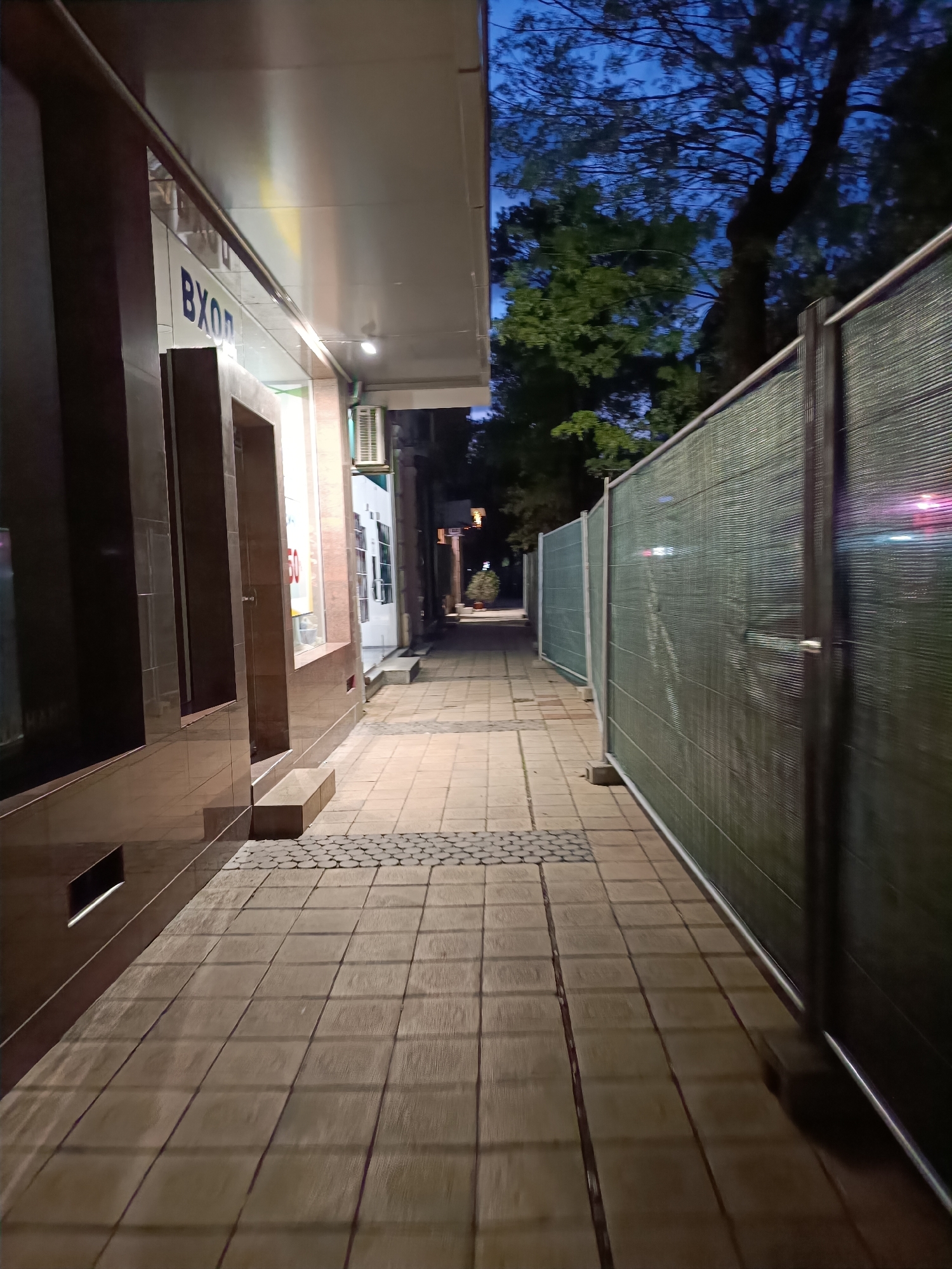 This media file captures a quiet urban alleyway or sidewalk in Pleven, Bulgaria, at dusk or nighttime. The path is deserted, with no people or activity visible.
On the left, a building facade runs alongside the path, featuring dark, reflective tiled or stone panels and large recessed window areas. A bright sign above the entrance displays the partially visible Cyrillic word "ВХОД" (Vkhod, meaning "Entrance") in blue letters on a white background. Further down the building, an air conditioning unit is mounted on the wall. The underside of a light-colored overhang or canopy extends above the path from the building.
To the right, a long temporary fence or hoarding delineates the other side of the path. This barrier consists of dark green mesh fabric stretched over a framework of metal poles and beams, suggesting a construction site or restricted area beyond. The fence recedes into the distance, indicating the path continues for some length.
The ground is paved with light-colored square tiles, interspersed with a small section of dark, circular cobblestones near the building's edge. In the background, dark tree canopies are silhouetted against a deep blue, twilight sky, hinting at natural elements beyond the immediate urban structures. Distant artificial lights are visible at the far end of the path. The overall atmosphere is tranquil and utilitarian.