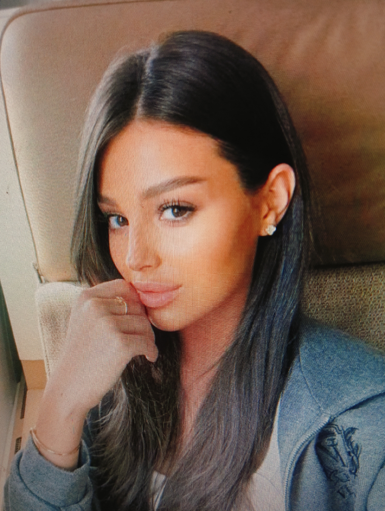 A woman with dark hair and fair skin is positioned in the foreground of the image, with her face turned slightly towards the viewer. She is wearing a light-colored jacket with blue accents and a white top underneath. A delicate bracelet adorns her left wrist, and a ring is visible on her left ring finger. Her right ear is adorned with a stud earring. The background appears to be an indoor setting with textured upholstery, possibly a seat in a vehicle or a chair. The lighting is soft, highlighting her facial features and attire. The image is a close-up shot, focusing primarily on the woman's upper body and face. No specific location in Sluck, Belarus is identifiable from the provided image.