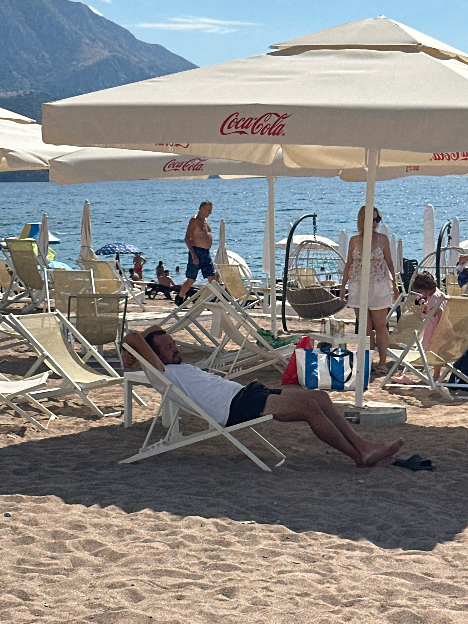In the city of Boreti, Montenegro, this image captures a sunny day on a beach with a man relaxing in a lounge chair. He is wearing a white t-shirt and dark shorts, with his arms behind his head. Several other lounge chairs are visible, some empty and some occupied by people. In the background, the clear blue sea stretches out, with a few boats in the distance. Beach umbrellas, some bearing the Coca-Cola logo, provide shade. A woman in a floral top and white shorts stands near a hanging chair, while a young child in a pink dress is nearby. The sand is light brown and textured, with footprints and shadows cast by the furniture. The overall scene depicts a typical beach resort atmosphere.