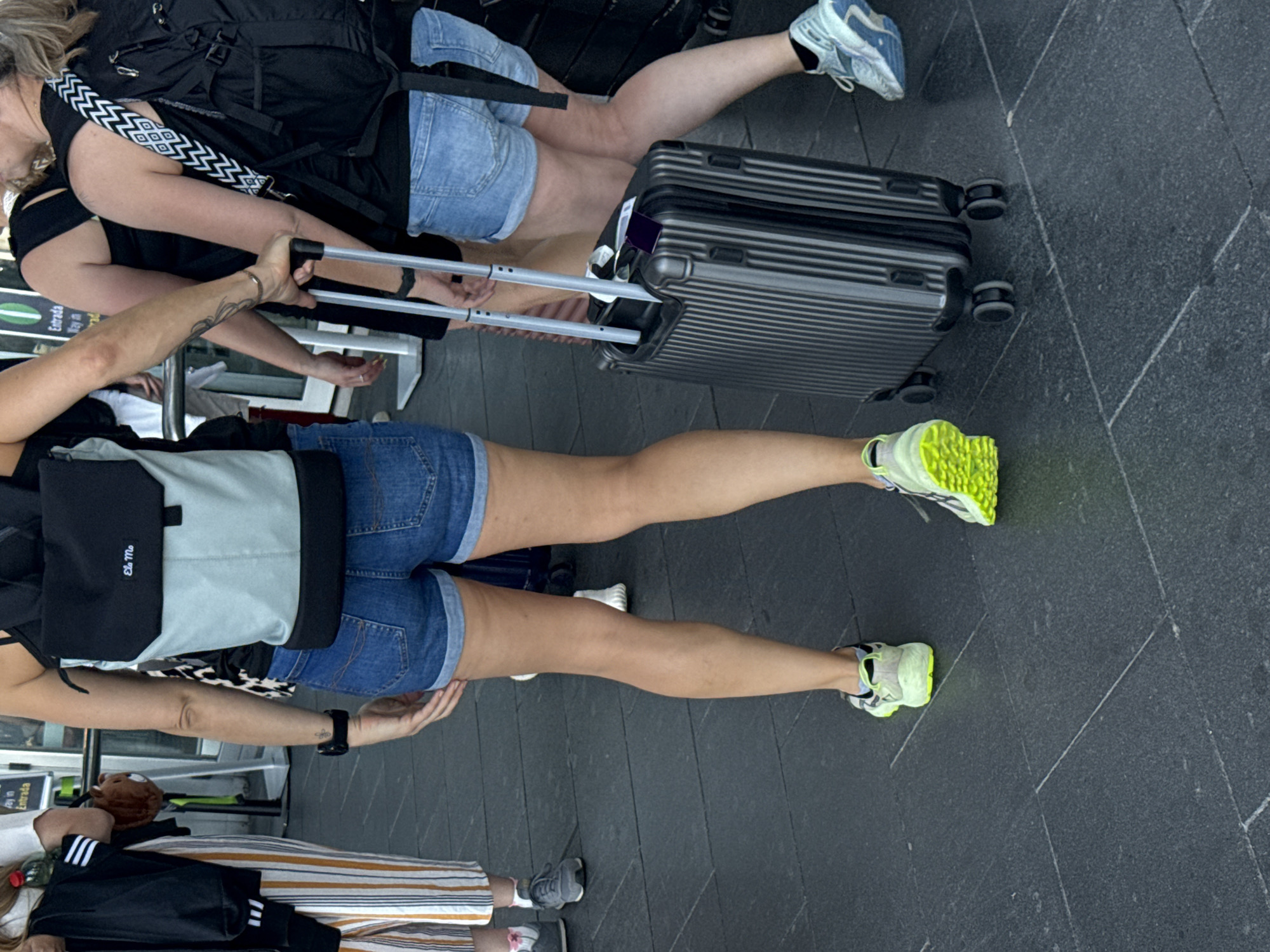 A group of people are walking on a dark gray paved surface, likely outside a terminal or transportation hub. The person in the foreground, facing away from the camera, is wearing denim shorts, a backpack, and bright neon yellow athletic shoes. They are pulling a dark gray, ribbed wheeled suitcase with a telescoping handle. To their right, another person is also walking away, wearing denim shorts and light blue athletic shoes, and carrying a black backpack with a decorative strap. To the left of the foreground person, another individual is walking towards the camera, wearing striped pants and athletic shoes. The background shows parts of a building with signage, including "Entrada" and "Salida," suggesting a public entrance or exit. The scene appears to be in El Prat de Llobregat, Spain, given the context of the image.