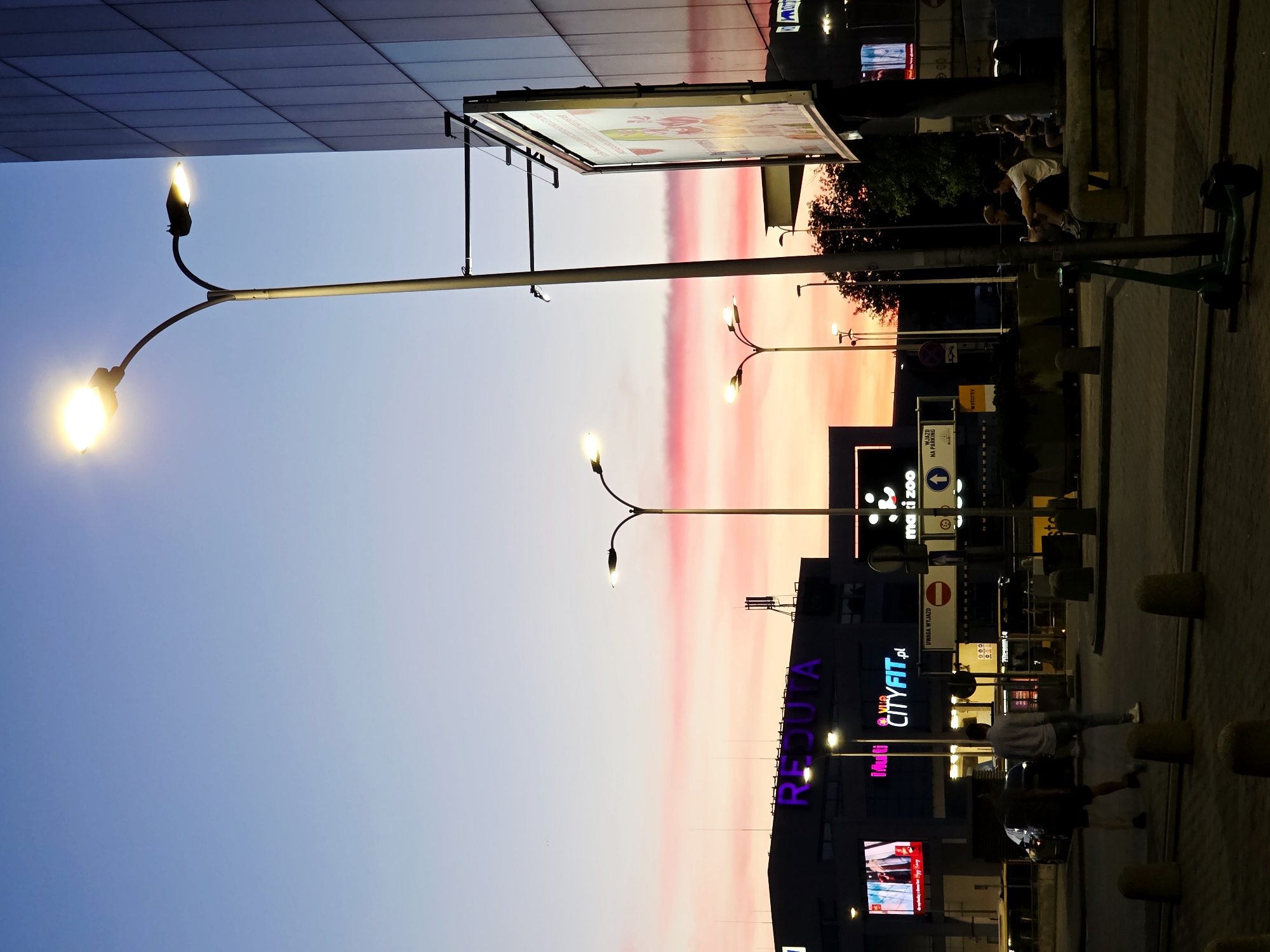 An outdoor street-level view captures a city scene at dusk with a vibrant sky displaying hues of pink, orange, and blue. A large, modern building with a reflective facade stands to the right, its vertical lines accentuated by the dimming light. In the foreground, a tall streetlamp with two illuminated bulbs casts a bright glow. To the left, a shopping center or complex displays prominent signage, including "REDUTA" in large purple letters and other brand names like "Multi" and "CITYFIT.pl" on illuminated screens. Several other streetlights are visible, some lit and others off. A billboard is mounted on the side of the large building.
On the ground, a paved area with circular concrete bollards suggests a pedestrian walkway or public space. Two figures are walking away from the camera in the middle ground, one in a light-colored long-sleeved shirt and jeans, the other in darker clothing. Further to the right, a group of individuals is seated on a low wall. A green electric scooter is parked near the streetlamp in the foreground. The overall atmosphere suggests a transition from day to night in a commercial or urban hub.