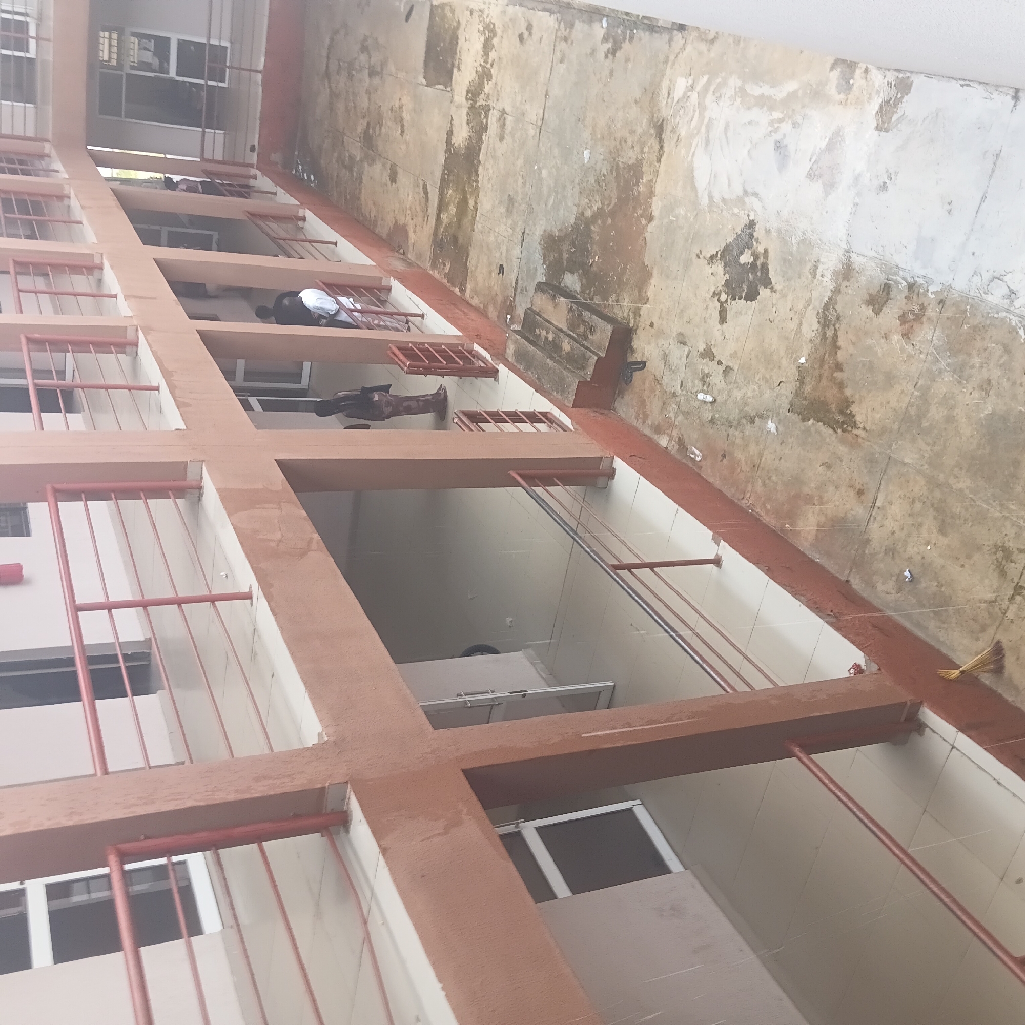 The image shows an outdoor corridor of a building with multiple levels. The corridor has a reddish-brown painted railing and pillars. Several people are visible, some lying down on what appear to be beds or mats in the open rooms or balconies of the building. One person is dressed in white and pink, another in black. The ground outside the building is paved and appears weathered and dirty, with scattered debris including plastic bottles and paper. A broom with a straw head leans against a pillar. The lighting suggests it is daytime, and the setting appears to be a residential or dormitory-like structure. The architecture suggests a warm climate. The image is taken from a high angle, looking down into the corridor. There is no visible text in the image.