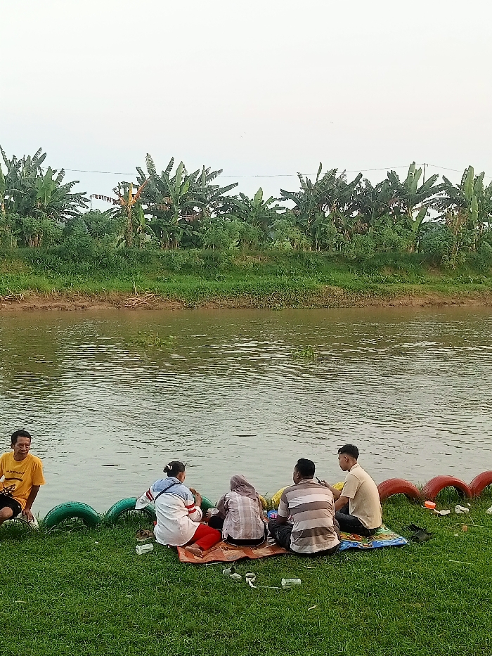 The image shows a group of six people sitting on a grassy bank next to a river. The river is calm and reflects the cloudy sky. There are banana trees growing along the opposite bank. The people are sitting in a circle on a yellow blanket. Three of them are facing the river, and the others are looking at the river. The people are wearing casual clothing. Some are sitting in front of what appears to be old tires that are now used as a barrier. The scene is located at coordinates (latitude: -6.758, longitude: 111.075) in the city of Jakenan, Indonesia.