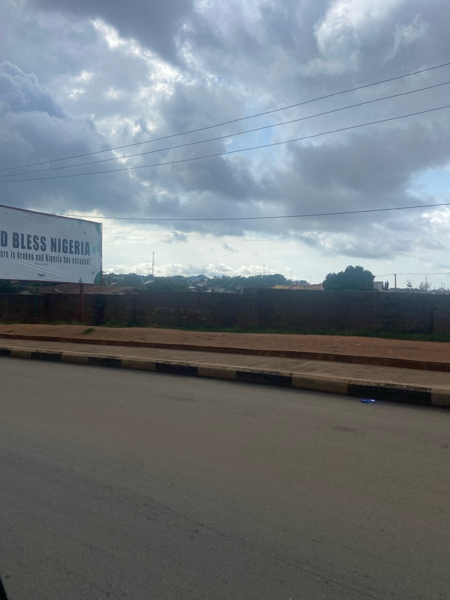 A wide shot captures a cloudy sky over a street scene in Jos, Nigeria. A large billboard displays "GOD BLESS NIGERIA" in bold lettering, with smaller text below it reading, "The snare is broken and Nigeria has escaped!" and "Psalm 124:7". Adjacent to the billboard, a dark brown brick wall stretches across the frame, separating a dirt path from a residential area with trees and buildings in the background. Power lines run horizontally across the sky. The foreground shows a paved road with a concrete curb and a narrow dirt shoulder. A small, bright blue object lies on the road near the curb. The overall atmosphere is overcast and somewhat muted, suggesting a possibly humid day.
