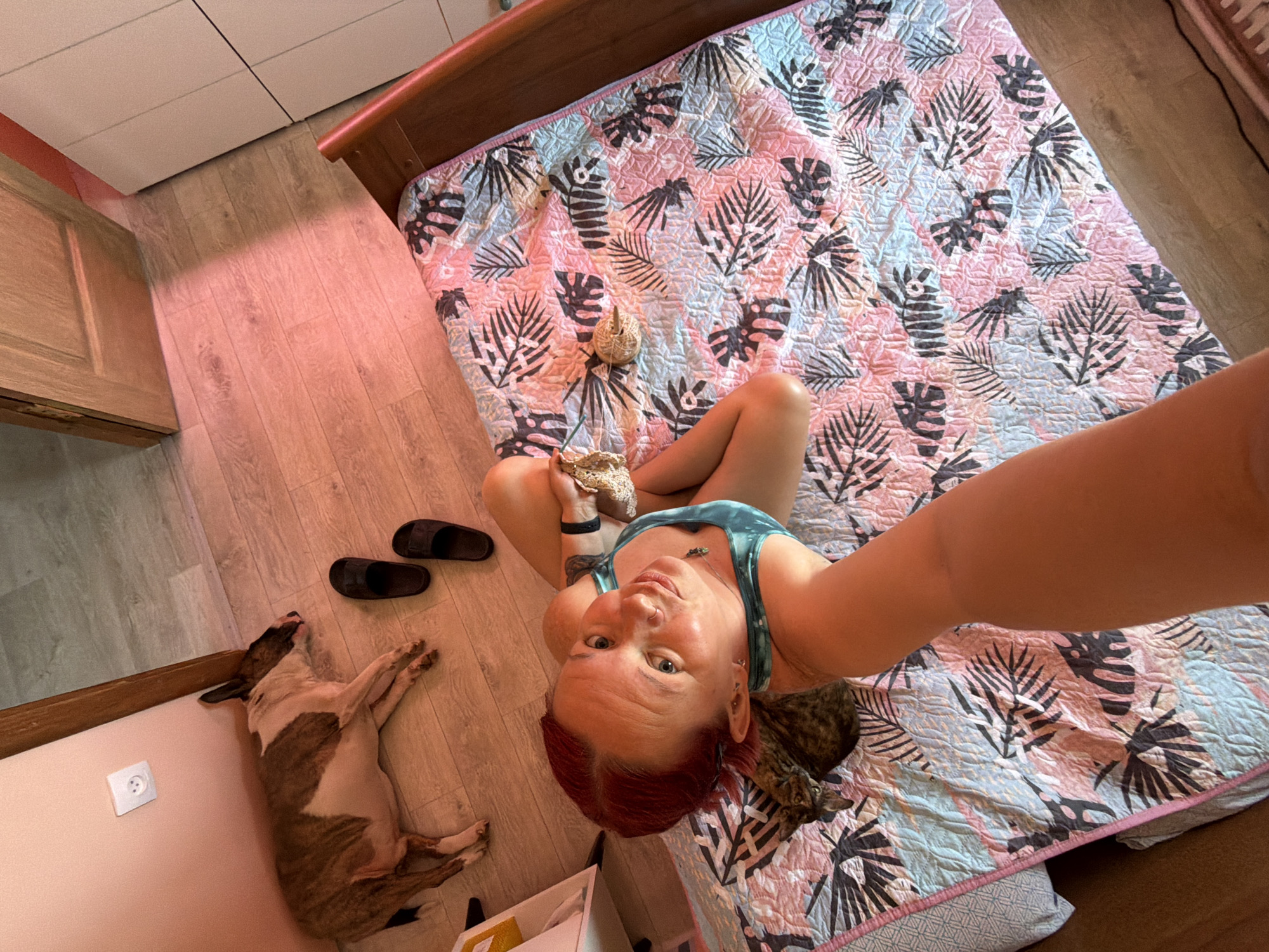 An overhead selfie shows a woman sitting cross-legged on a bed covered with a tropical-patterned quilt. She is holding a ball of yarn and crocheting with a needle. She has red hair, fair skin, and is wearing a blue tank top. A tabby cat lies near her on the bed. Near the bed on a hardwood floor, a brindle and white dog sleeps near a pair of black sandals. A white electrical outlet is visible on the wall near the dog. The room contains wooden furniture and is lit with a reddish glow. The image was taken in Chaikovsky, Russia.