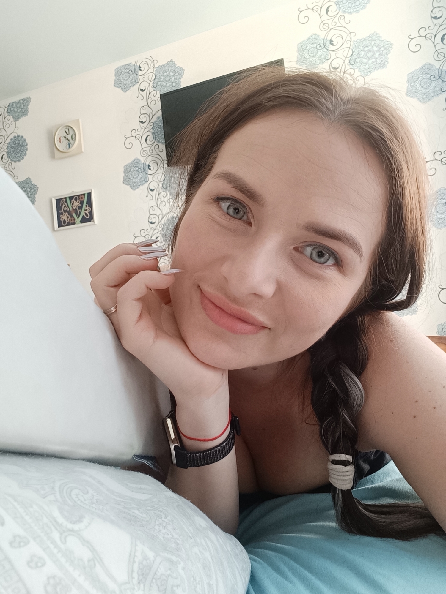 A woman with light skin, light blue eyes, and brown hair braided on one side is lying on a pillow. She has a watch on her left wrist and is wearing a ring on her left hand. The background consists of a wall with floral wallpaper, a clock, a framed picture, and a television. Based on the surrounding information, this photo was taken in the city of Chaikovsky, Russia.