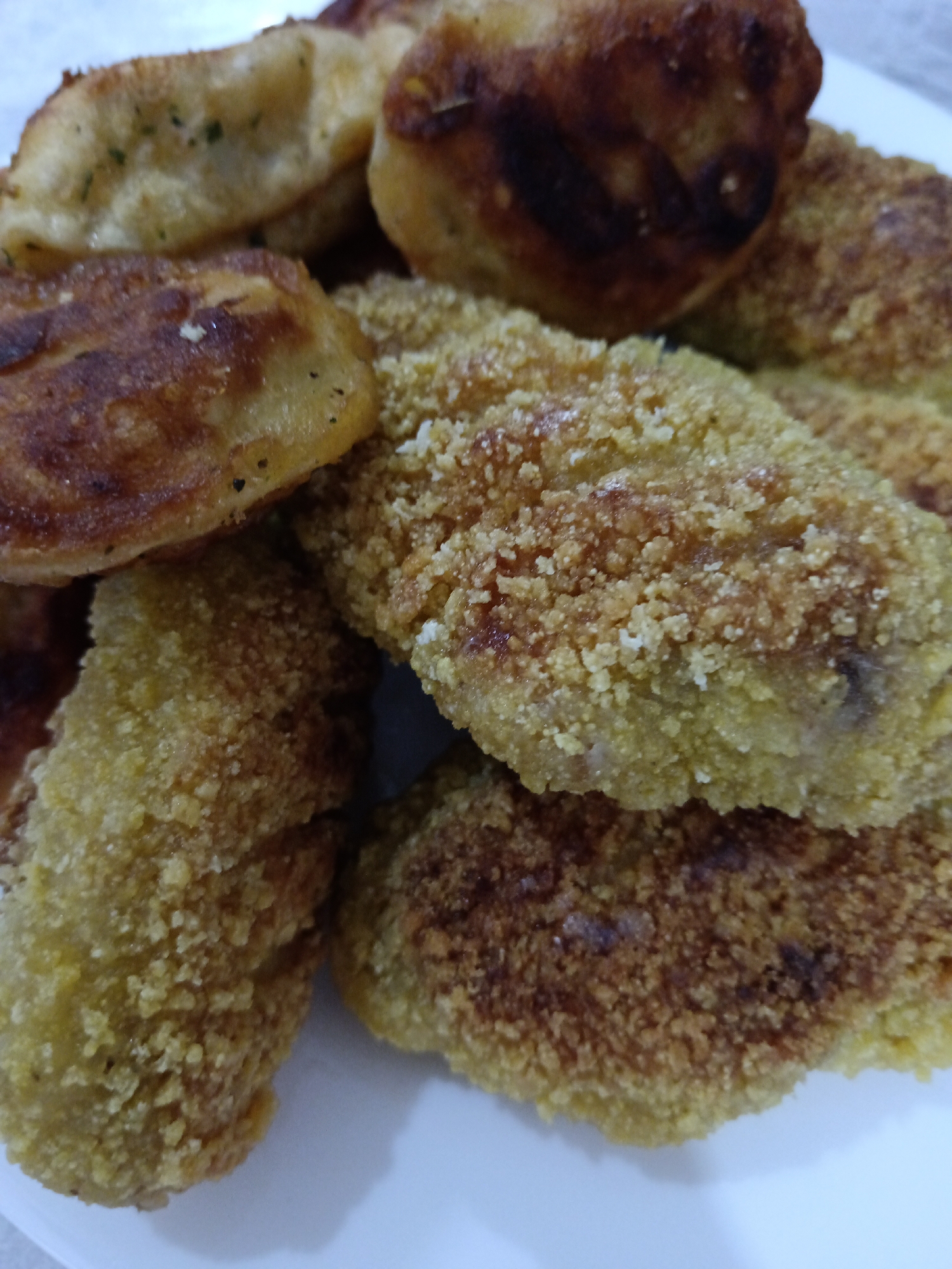 The image displays a close-up, overhead view of a white plate filled with several pieces of fried food. The food appears to be some type of breaded cutlets or nuggets, with a golden-brown, crispy exterior. Some pieces show darker, more heavily browned areas, suggesting they were fried longer or at a higher temperature. The texture of the breading is coarse, with visible crumbs. The food is piled on top of each other, creating a dense arrangement. No specific location in Sorochinsk, Russia, is depicted or implied by the image content. The focus is entirely on the food itself.