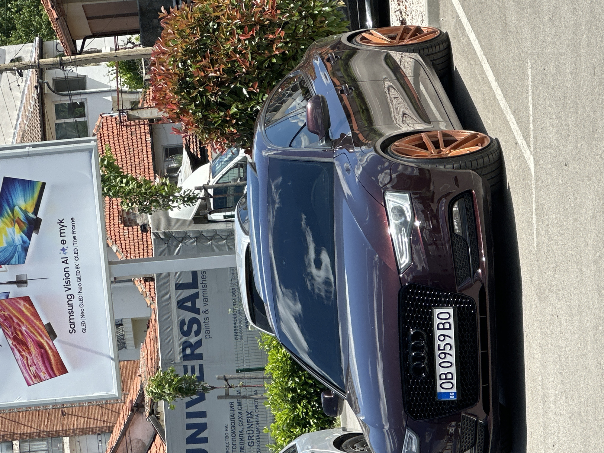 A maroon-colored Audi car is parked on a paved surface in front of a building in Unknown, Unknown. The car has custom bronze-colored rims and a Bulgarian license plate that reads "OB 0959 BO". A large billboard advertising Samsung Vision AI and Universal paints & varnishes is visible on the left, and some greenery is present between the car and the billboard. Other buildings with red-tiled roofs and a white van are visible in the background. The photo was taken in daylight under sunny weather conditions, with clouds reflected in the car's windows.