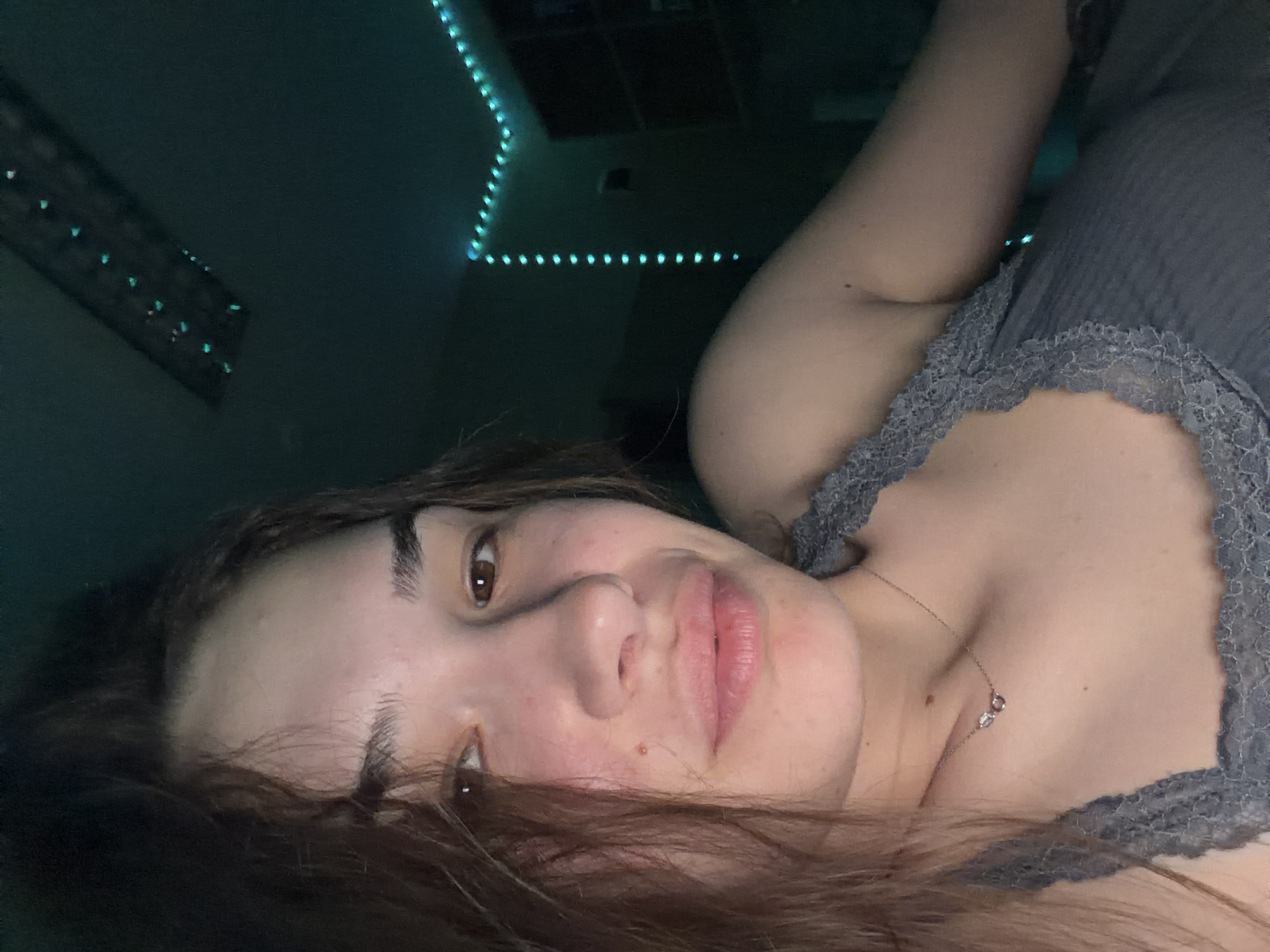 A young woman is depicted in a close-up, tilted shot, likely a selfie, taken indoors at night or in the evening in Sofia, Bulgaria. She is lying down, with her head resting on a surface, looking directly towards the camera. Her dark brown hair cascades around her face, partially obscuring her left side. She has brown eyes and a neutral to slightly smiling expression. Her fair skin shows some natural redness on her cheeks and forehead.
She is wearing a grey top with delicate lace trim along the neckline. A thin silver necklace with two small charms hangs around her neck, resting against her chest.
The background is dark and features prominent blue-green LED strip lights outlining a corner of a room, creating an atmospheric glow. Another section of similar lights or a reflective surface is visible further in the dark background to the left. The lighting is predominantly artificial, casting cool tones throughout the scene. No visible text is present in the image.