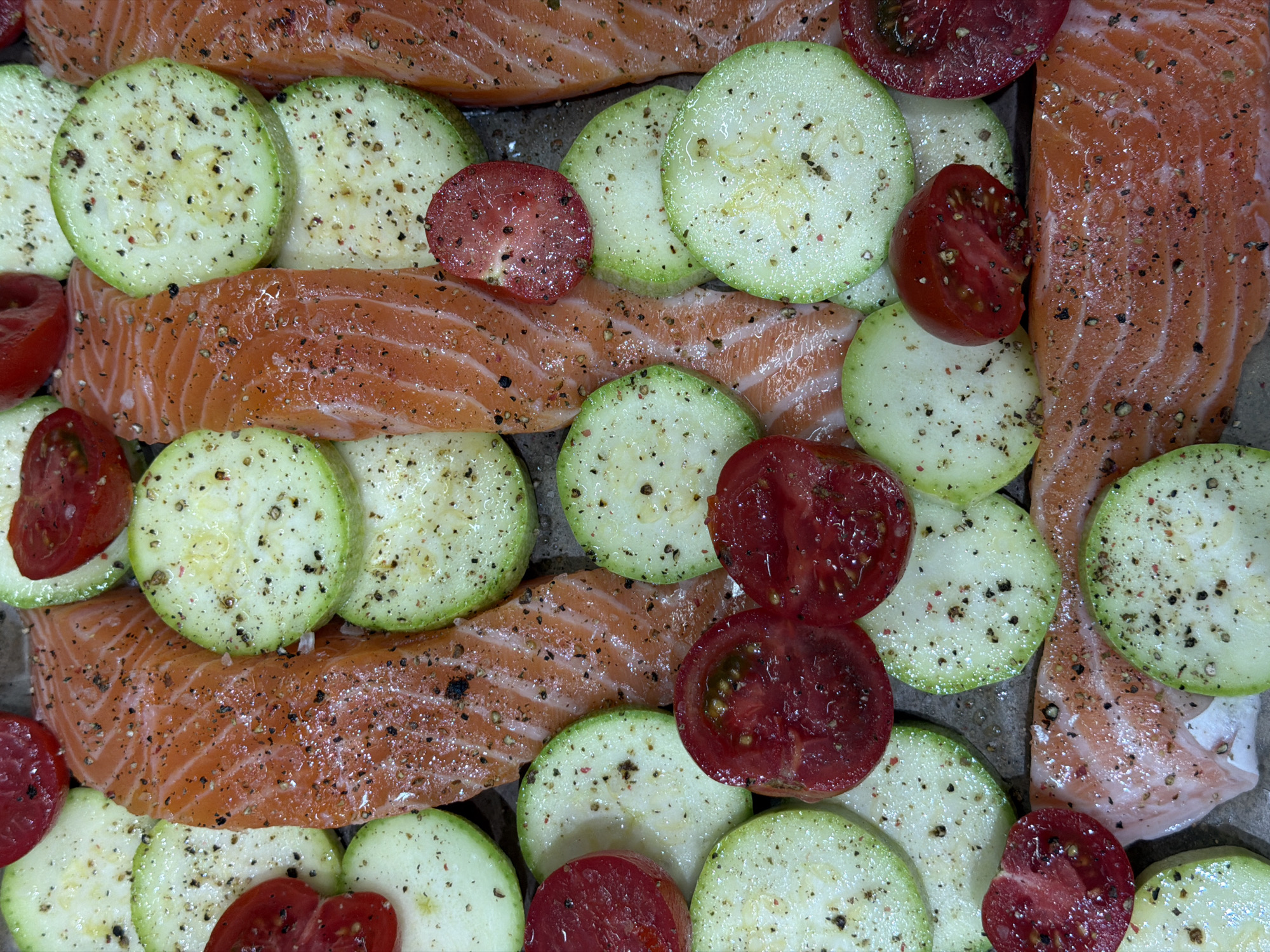 A close-up, high-angle shot reveals raw salmon filets intermixed with slices of zucchini and tomatoes, all seasoned with what appears to be black pepper. The food items are placed on a gray, slightly textured surface. No people or interactions are visible. The environment is domestic, seemingly inside a kitchen setting given the context of food preparation. There is no discernible location within the city of Moscow, Russia evident in the image.