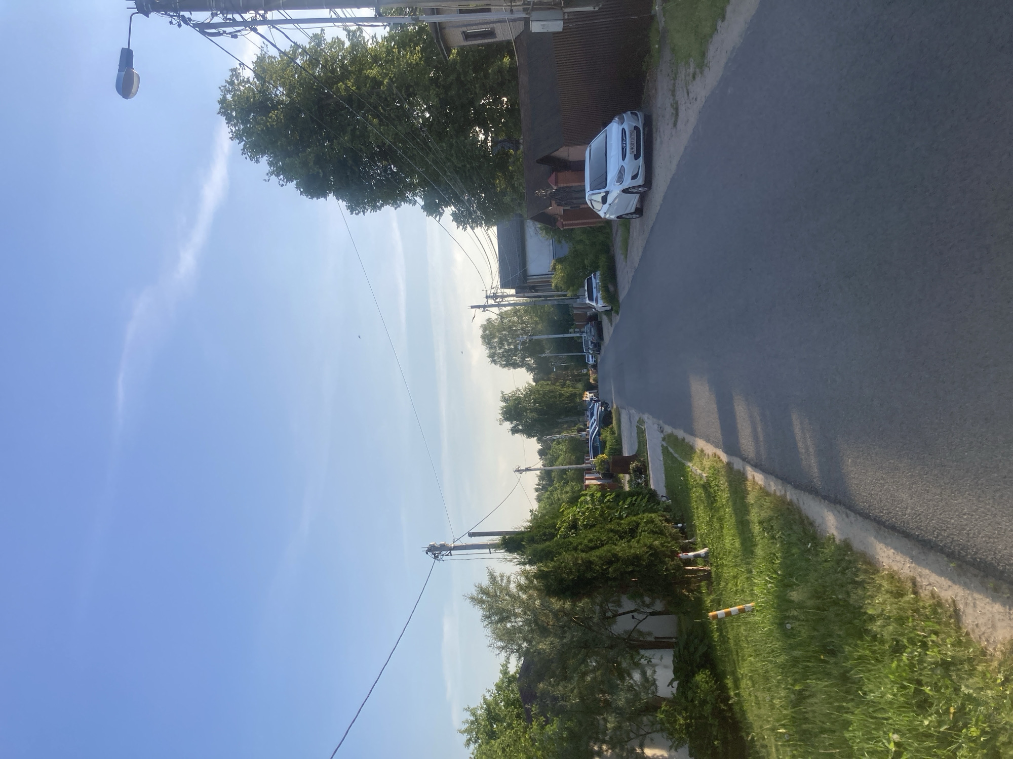 The image depicts a residential street scene in Novoaleksandrovo, Russia, captured under a clear, light blue sky with faint, wispy clouds visible in the upper right quadrant. The sun, positioned out of frame to the upper right, casts long shadows across the asphalt road, indicating late afternoon or early evening.
The asphalt road, dark grey and without markings, extends from the foreground into the background, narrowing slightly.
On the left side of the road, a strip of unpaved ground with green grass and some dirt runs parallel to the asphalt. A small, vertically oriented pole with alternating orange and white stripes is visible in this grassy area in the foreground. Behind this strip, a variety of vegetation, including manicured evergreen hedges and deciduous trees, lines the street. Utility poles with multiple overhead wires are present along this side, receding into the distance. Portions of residential fences and structures are discernible through the foliage.
On the right side of the road, a strip of dirt and sparse grass separates the pavement from a prominent brown fence constructed of vertical wooden slats. A white four-door Hyundai Accent sedan is parked on this dirt strip, parallel to the fence, facing away from the camera. The vehicle's rear license plate displays "X 903 TO 190". Further down the street, multiple other vehicles are parked. A tall utility pole, heavily laden with numerous black wires, stands near the fence in the mid-ground, extending upwards into the sky. A blue-housed streetlight fixture is attached to this pole. Beyond the fence and utility pole, a large, dense deciduous tree with green foliage dominates the right mid-ground, and parts of additional residential buildings are visible.
No individuals or active traffic are visible in the scene. The environment appears to be a quiet suburban or semi-rural residential area.