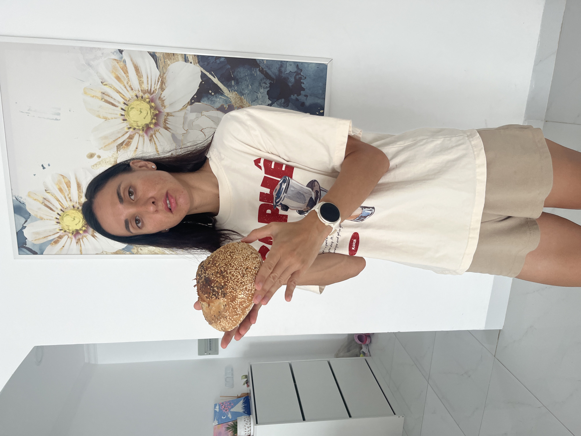 A woman is holding a sesame seed-covered loaf of bread in her hands. She is wearing a white t-shirt, beige shorts, and a white smartwatch. Behind her is a framed floral print on a white wall. The floor is white tile. In the lower left corner are white cabinets. This is in Bac Nha Trang Ward, Vietnam.