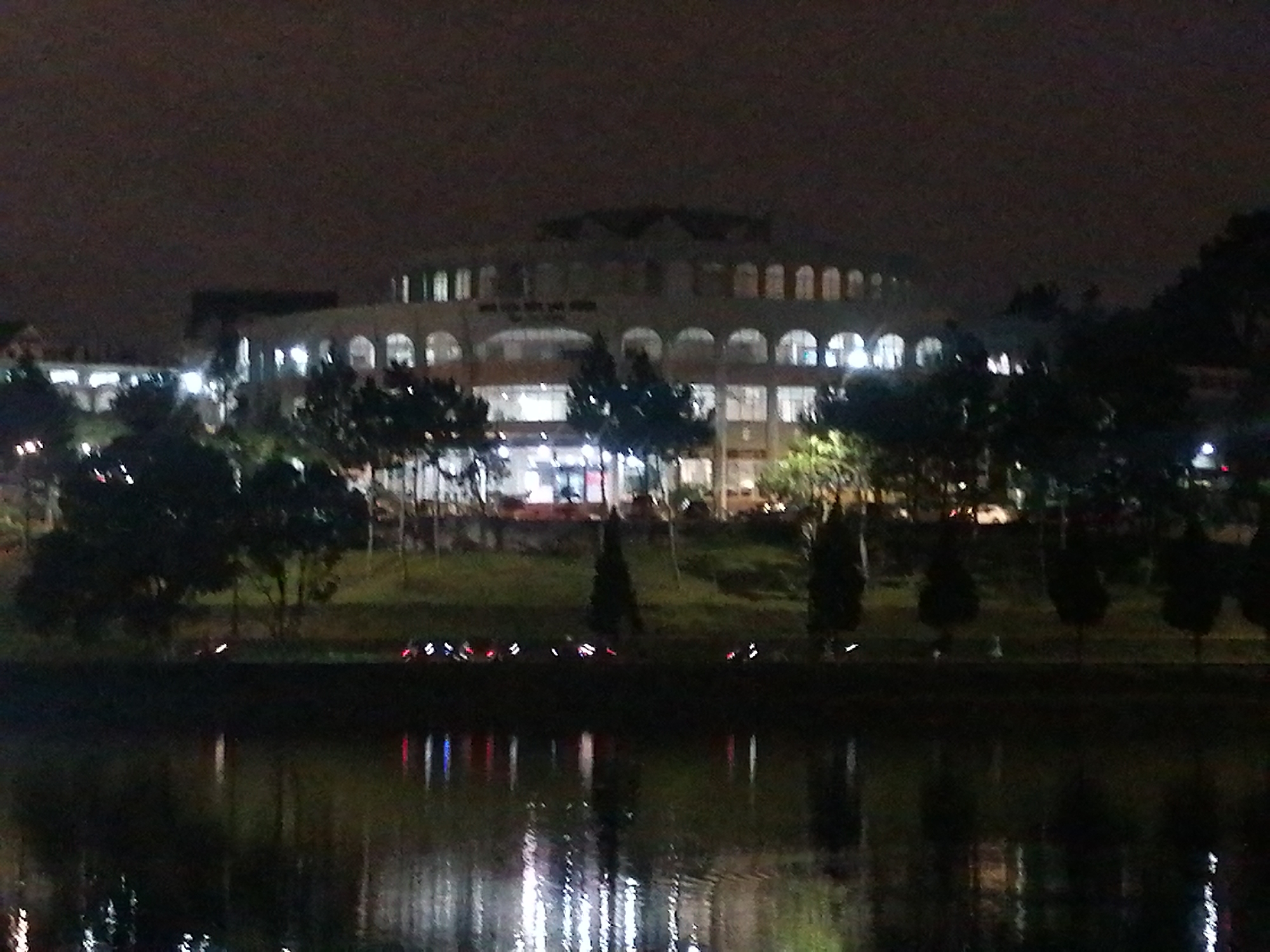 At night, a three-story building with a rounded roof and arched windows is viewed across a body of water. The building is lit from within, and its illumination reflects in the water. Trees surround the building on either side. The location appears to be in Da Lat, Vietnam. The sky is dark. The photo has low light and appears to be taken from a distance.