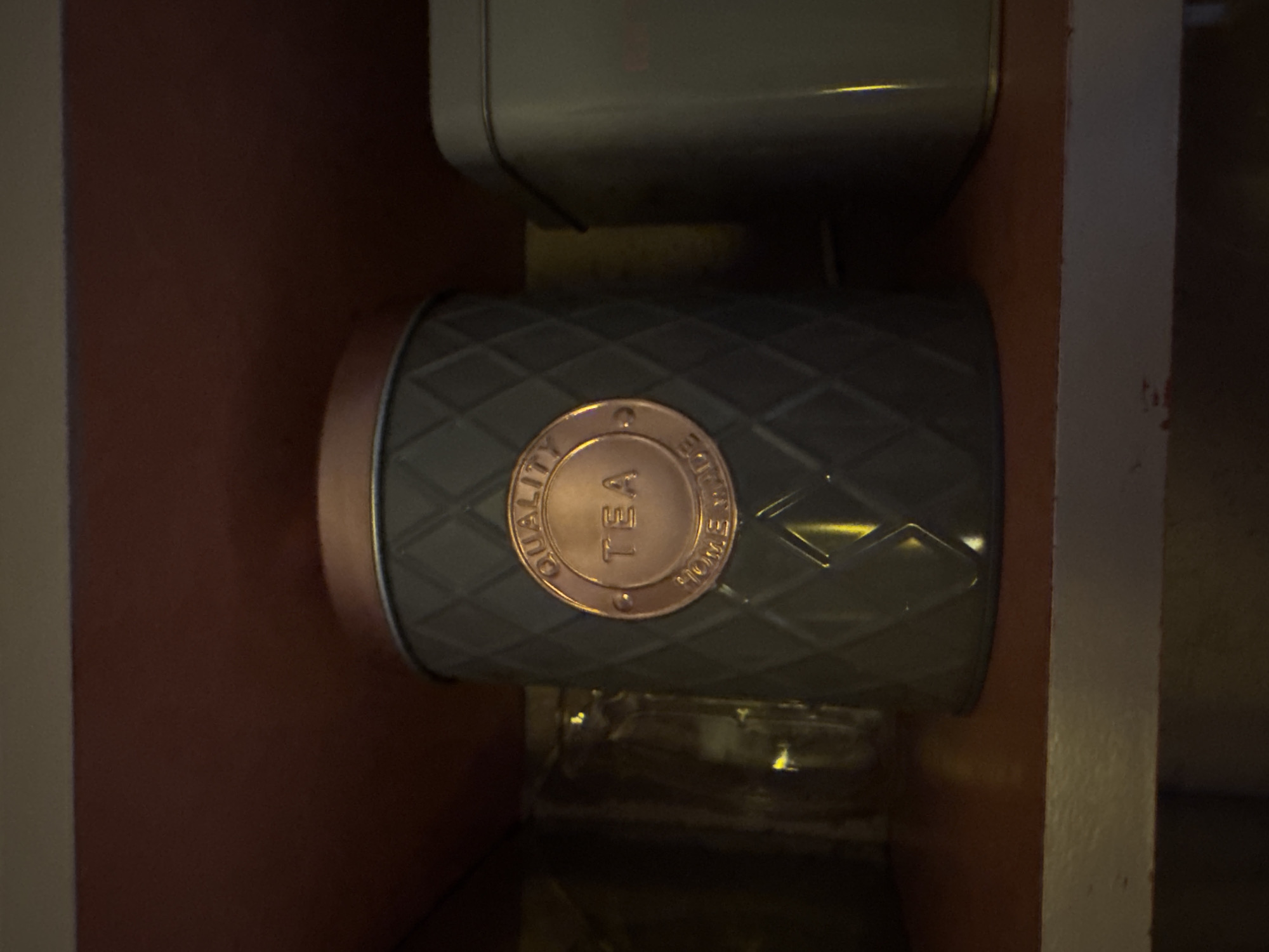A close-up, eye-level shot captures a dark gray, textured tin canister with a copper lid and a copper circular emblem on its front. The emblem is engraved with the words "QUALITY," "TEA," and "HOMEMADE." The canister sits on a dark wooden shelf, beneath a reddish-brown surface. To the right of the central canister is another, larger tin with a light gray lid. The overall lighting is dim, with some bright yellow highlights reflecting off the surfaces. The background is dark and out of focus, suggesting an indoor, possibly domestic, setting.