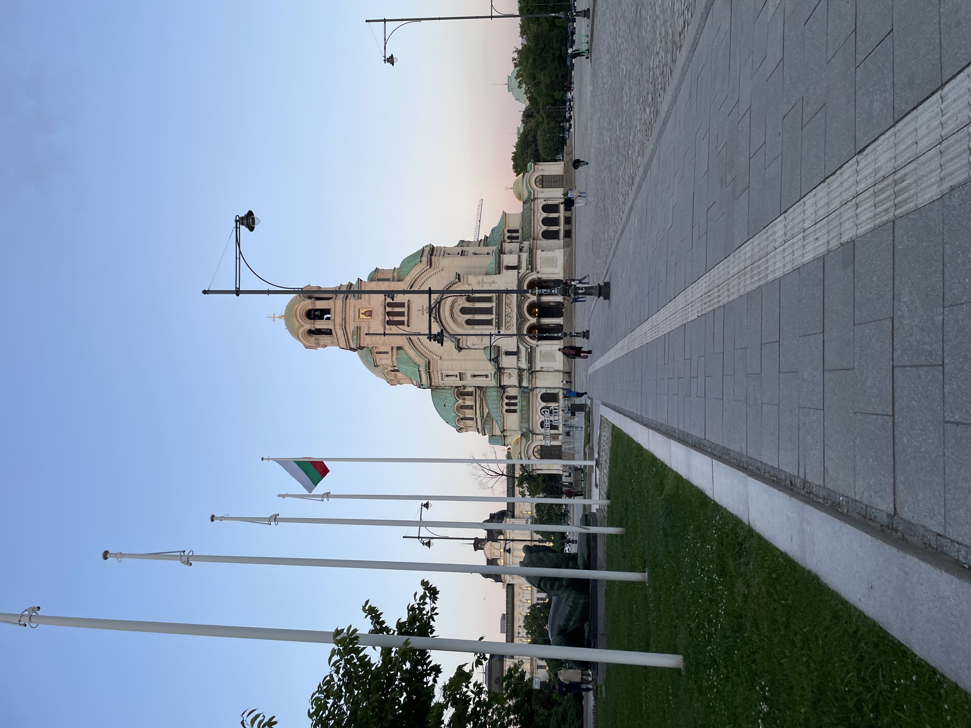 The image shows the Alexander Nevsky Cathedral in Sofia, Bulgaria. The view is taken from a low angle, positioned near the sidewalk. In the foreground, there are flag poles, some without flags. The Bulgarian flag is visible on one of the poles. Green grass is visible between the poles and the sidewalk. The sidewalk is paved with grey tiles and features a tactile paving strip. Some people are seen in the distance on the sidewalk near the Cathedral. The sky is a light blue color grading into pink near the horizon. Several street lamps are visible.