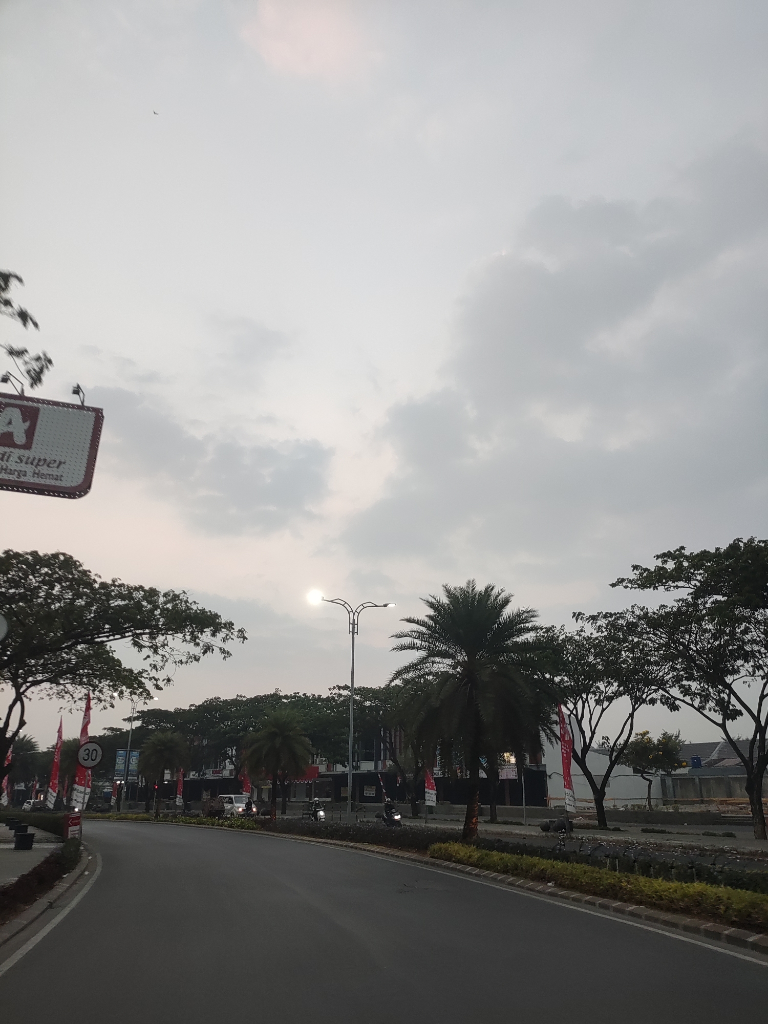 A street view captures a road curving to the left, lined with trees and palm trees on the right. The sky is overcast with muted light, and the sun is a bright, diffused circle near the horizon, partially obscured by clouds. A street lamp with a distinctive decorative design stands in the middle ground. On the left side of the road, a speed limit sign indicating "30" is visible, along with several red flags. A sign for "di super Harga Hemat" is also present. Buildings are visible in the background, suggesting an urban or suburban environment. Several motorcycles and a van are on the road, with some individuals riding the motorcycles. The scene appears to be in Telagaasih, Indonesia, likely during the late afternoon or early morning due to the lighting conditions.
