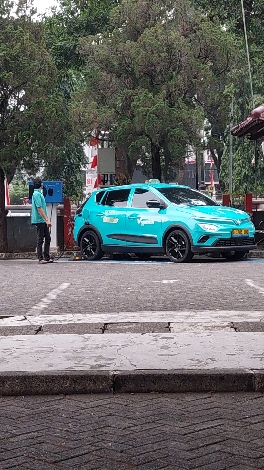 A turquoise electric vehicle, marked with "GREEN SM," is parked and plugged into a charging station on a street in Cibinong, Indonesia. A person wearing a light blue shirt and dark pants stands by the charging station, holding a charging cable connected to the vehicle. The car is a hatchback with black rims and tinted windows. In the background, there are trees, a building with a red-tiled roof, and what appears to be a flag. The charging station is blue and mounted on a pole. The scene is captured on a cloudy day.