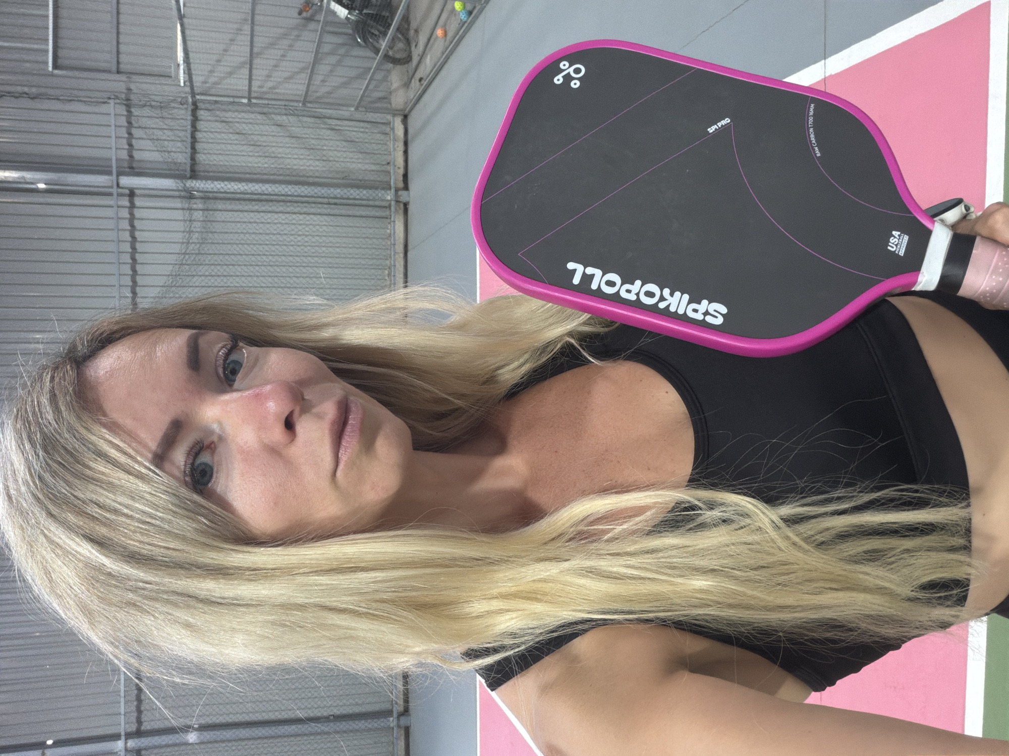 A blonde woman is taking a selfie in what appears to be an indoor pickleball court. She is wearing a black sports bra and holding a black and pink pickleball paddle. The paddle has the brand name "SPIKPOLL" printed on it. The background shows a chain-link fence, a concrete floor, and court markings. The lighting is artificial and appears to be from overhead. There are also some colorful balls scattered on the floor in the background. The woman has a neutral expression and is looking at the camera.