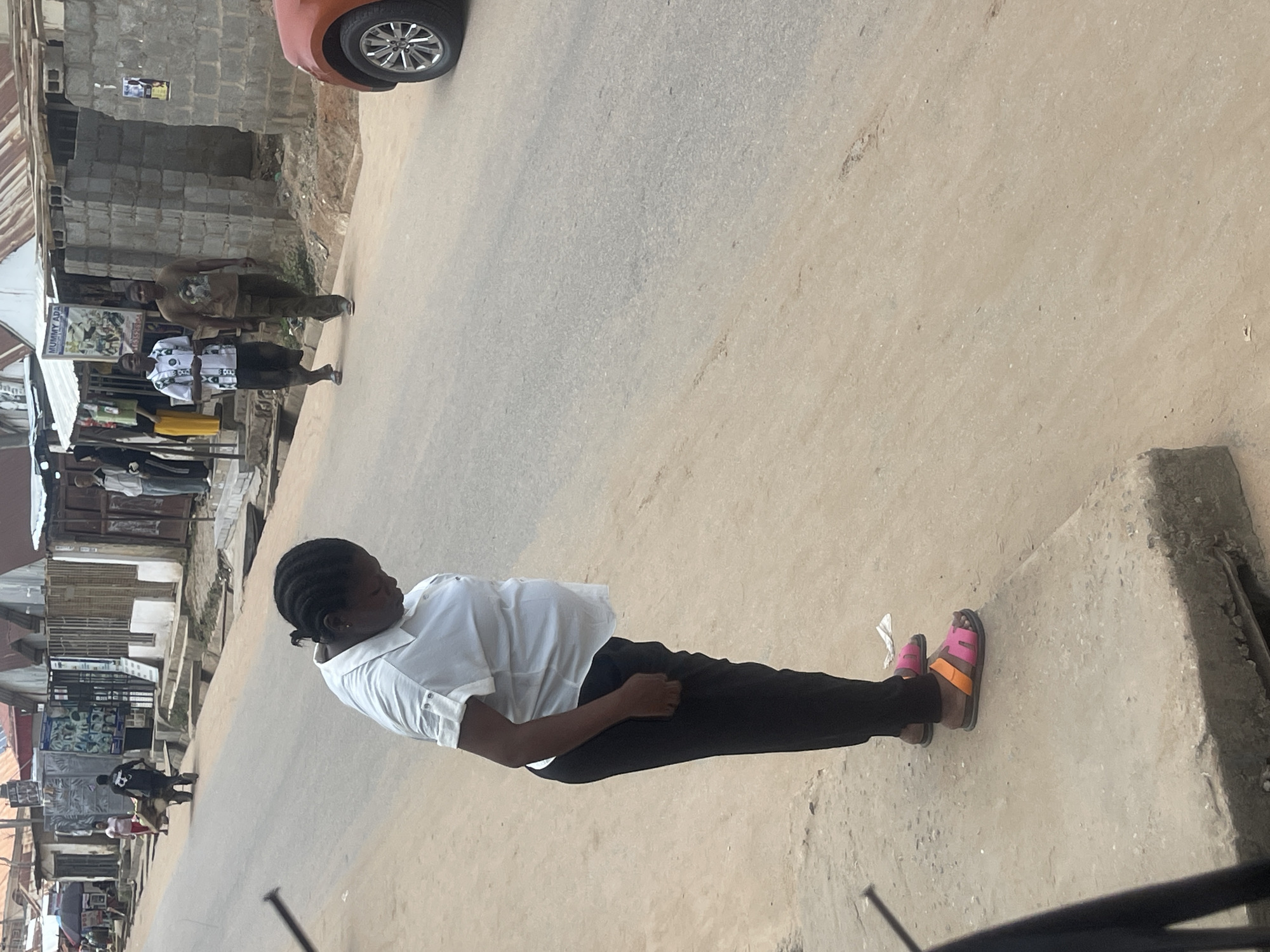 In Abuja, Nigeria, a Black woman stands on a concrete sidewalk, her body turned to the side, head bowed. She is wearing a white short-sleeved button-down shirt and black trousers. Her feet are adorned with pink and orange slide sandals. Behind her, on the street, two men walk away from the camera. The man on the left wears a patterned white and black shirt and dark trousers. The man on the right is dressed in a brown t-shirt and khaki trousers. Further in the background, other individuals and structures line the street, suggesting an urban environment. An orange vehicle is partially visible on the right side of the frame. The ground is a mixture of paved road and sandy earth.