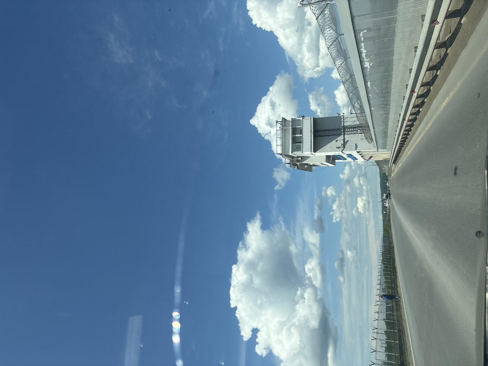 The image depicts a view from inside a vehicle driving along a road next to a large wall with barbed wire. The sky is blue with white cumulus clouds. Adjacent to the road, a gray concrete structure with an observation deck and a metal staircase stands against the sky. The location is likely a controlled or restricted area, suggested by the security measures like barbed wire and the imposing wall. The road appears to be paved and has a yellow line marking. Further down the road, light poles and some vegetation can be seen along the fence line. The scene is in daylight, and the angle suggests the image was taken from within a moving vehicle. The city of Chaikovsky, Russia, is specified as the location.