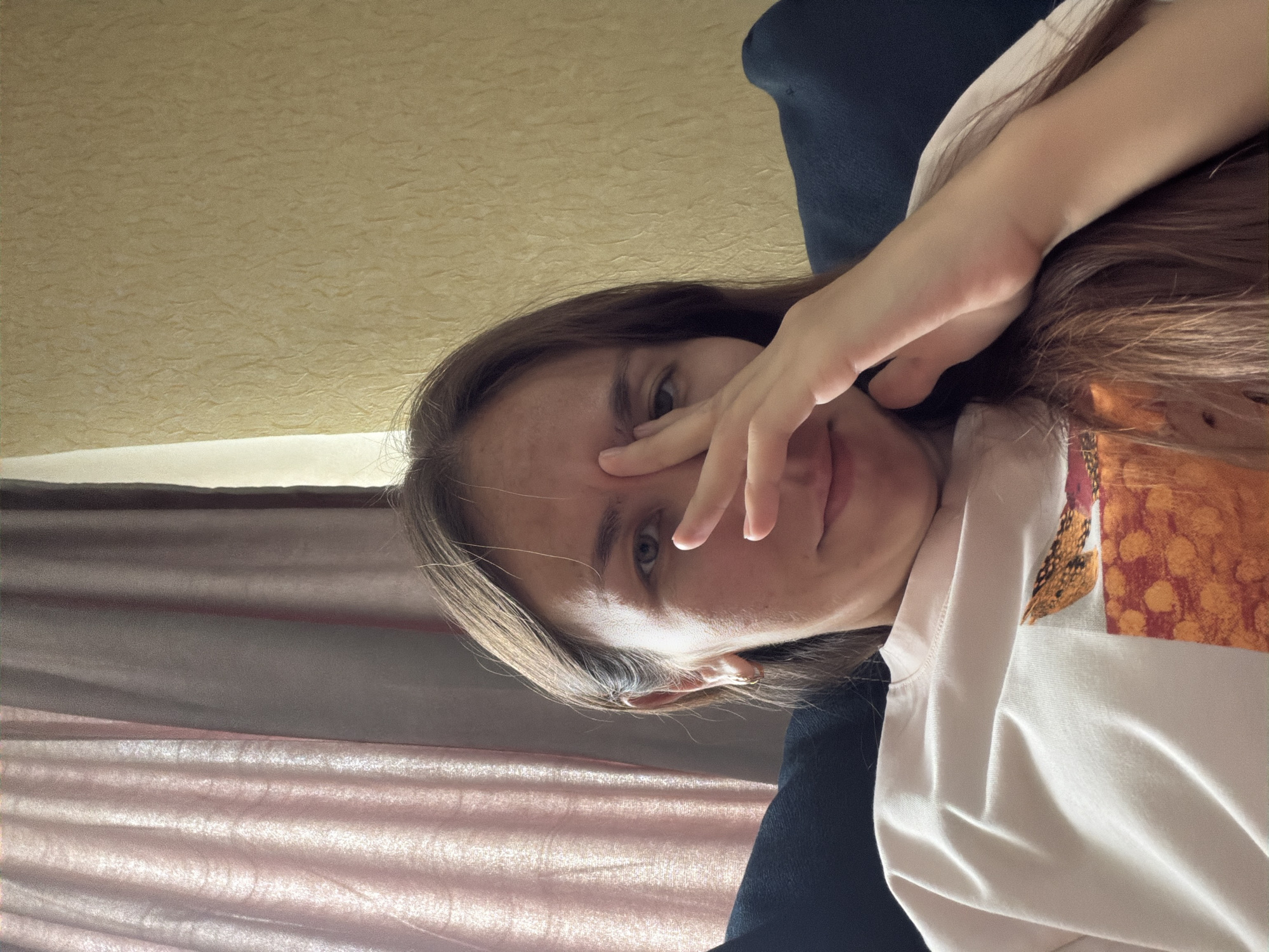 A young woman with long, brown hair is taking a selfie in a room. She is resting her head on her hand, with her fingers partially covering her eye and forehead. She is wearing a light pink t-shirt with a graphic print. The background includes a textured yellow wall and a curtain. The image appears to have been taken indoors, likely in a residential setting. There is no discernible information about the city of Tambov, Russia, or any specific location within it.