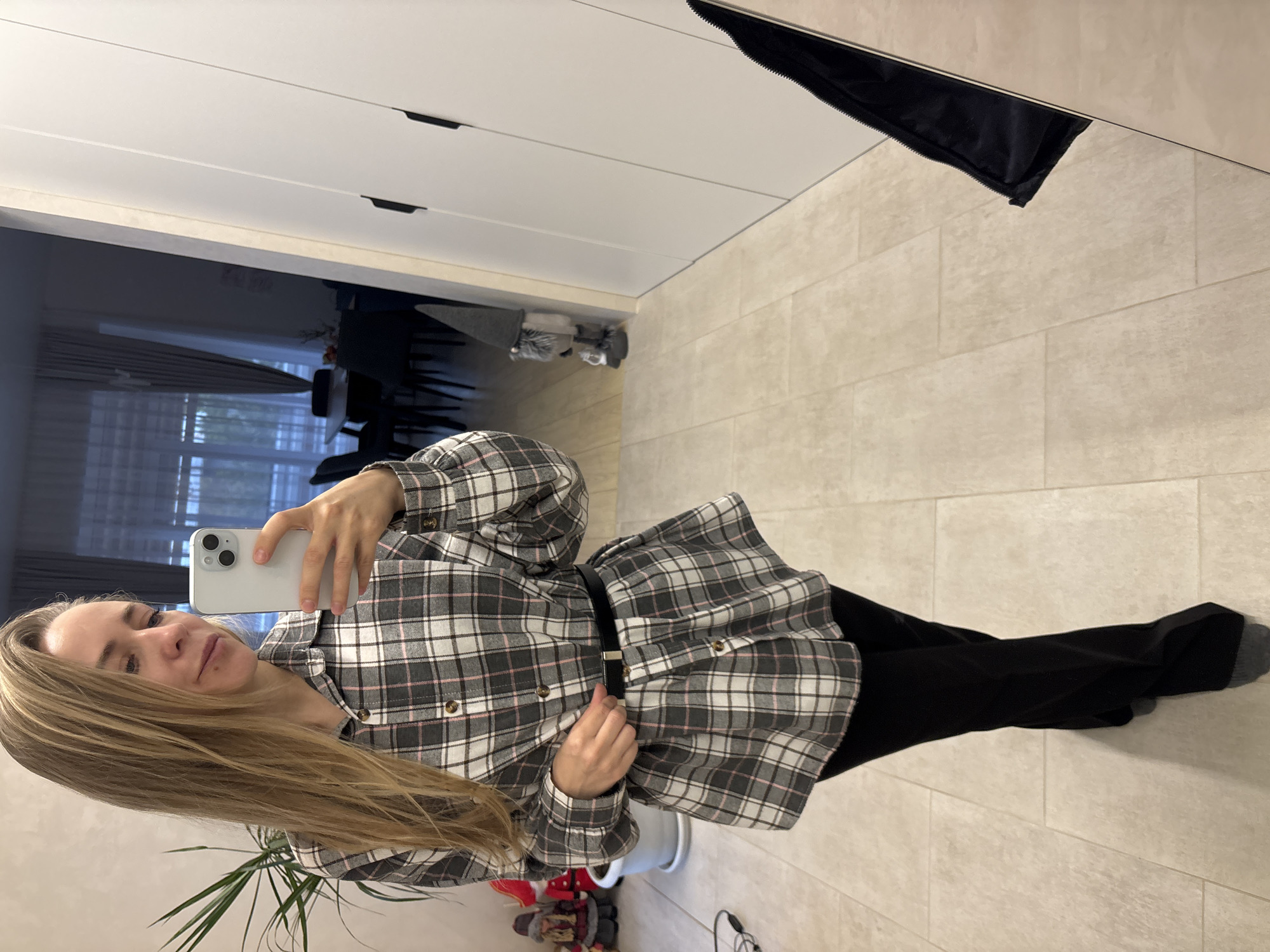 A woman is taking a selfie in what appears to be an indoor setting. She is holding a white smartphone, presumably to capture the image in a mirror. She is wearing a plaid, long-sleeved shirt cinched with a black belt and dark pants. The background includes a white wardrobe or closet with minimalist, black handles. The floor is tiled with large, light-colored tiles. A glimpse into another room reveals a dining area with chairs. There are plants in pots near her feet. It is impossible to determine the location in the city of Chaikovsky, Russia from the image.