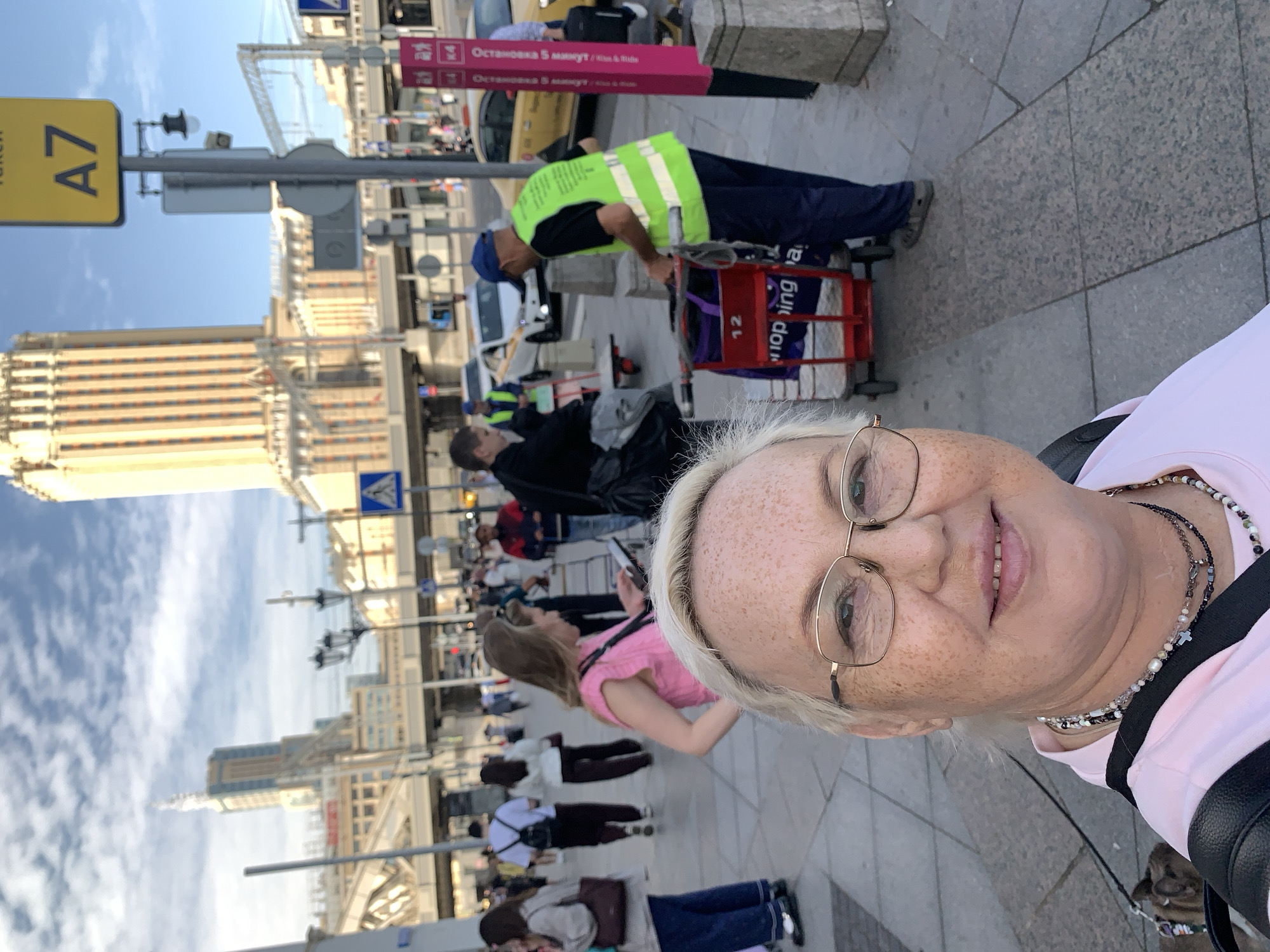 This is a selfie taken by a woman with blonde hair and glasses, wearing a pink t-shirt and necklaces. She is in Moscow, Russia, in front of the main building of Moscow State University, also known as MSU or Lomonosov University, a prominent Stalinist Empire-style skyscraper. In the background, other people are visible walking, and some are pushing carts. One man in a bright green vest is pushing a red cart with purple bags. A yellow taxi is also visible. The sky is blue with some clouds.