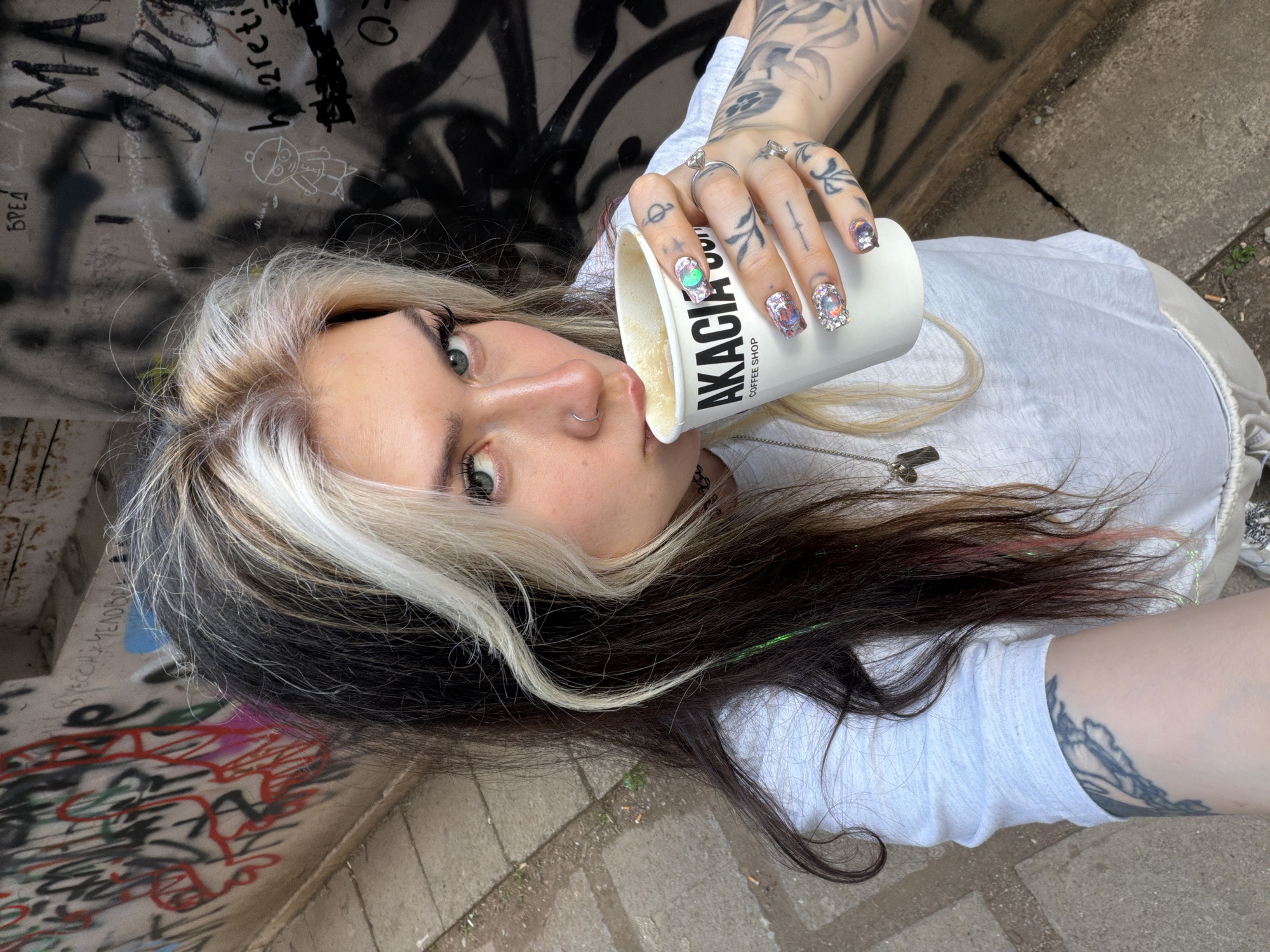 This image is a portrait of a young woman, likely a selfie, taken from a slightly overhead, tilted perspective. The woman is positioned on her side, looking directly at the camera with her light-colored eyes, possibly green or hazel. She has distinctive two-toned hair: dark brown or black with a prominent white or blonde streak at the front. A small silver hoop nose piercing is visible.
She is holding a white takeaway coffee cup to her lips, appearing to drink from it. The cup clearly displays the text "AKACIA COFFEE SHOP". Her right hand, holding the cup, is adorned with rings, including one featuring a large, clear gemstone. Her fingernails are elaborately decorated with large, sparkling, multi-colored gems. Tattoos are visible on her fingers and arms. She is wearing a light-colored (possibly white or light grey) t-shirt, and a silver necklace with a rectangular pendant hangs around her neck.
The setting is an urban outdoor environment in Budva, Montenegro, characterized by a heavily graffiti-covered concrete wall in the background. The graffiti includes various tags, scribbles, and a simple drawing of a figure. Some Cyrillic text, such as "БРЕД" (meaning "nonsense"), is visible among the markings. The ground is paved with concrete blocks. The lighting suggests it is daytime, possibly overcast or in a shaded area, without harsh direct sunlight. The overall atmosphere conveys a casual moment captured in a raw, street-art-filled urban landscape.