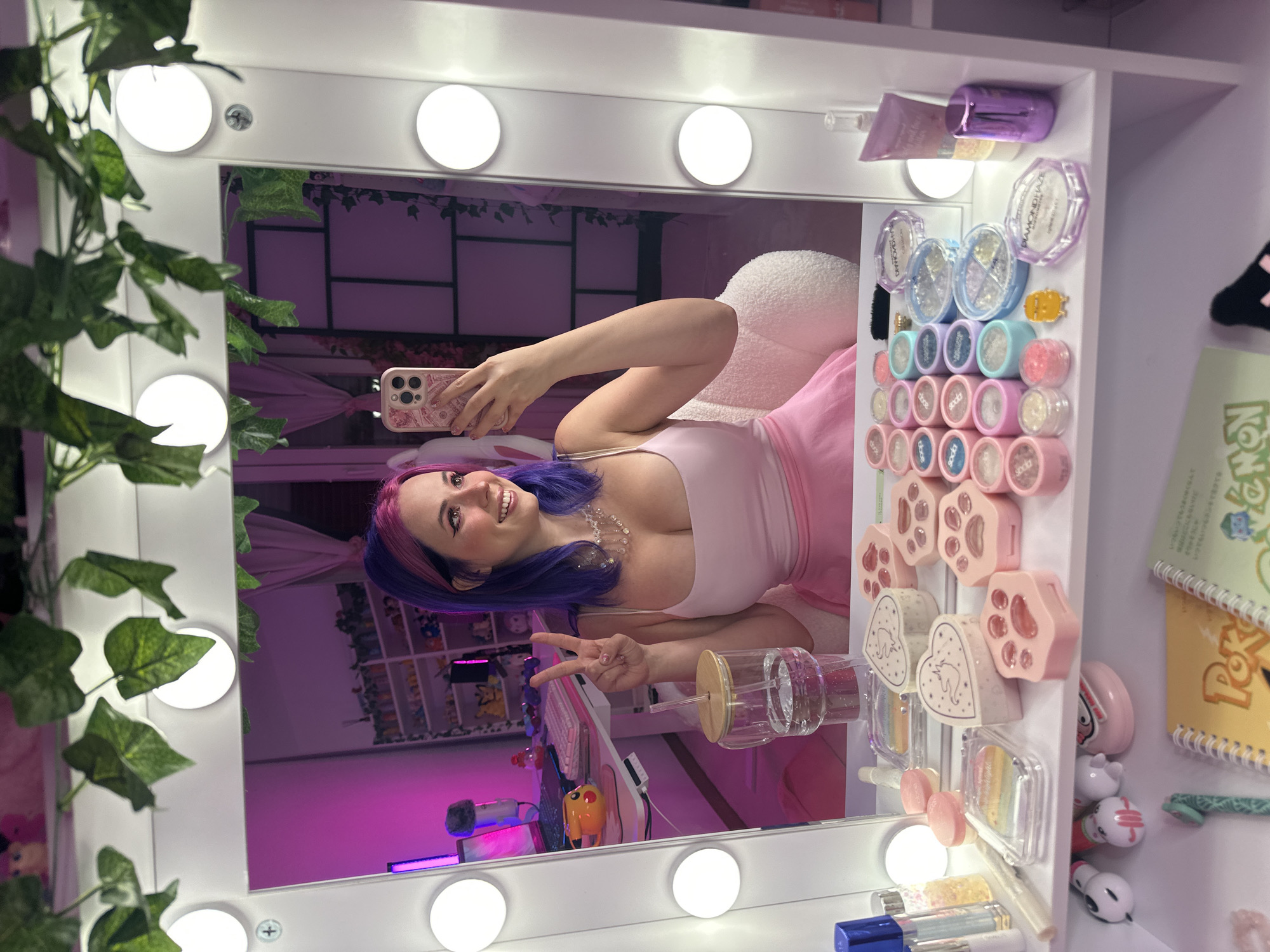 The image shows a young woman with purple and pink hair taking a selfie in a vanity mirror. She is wearing a pink spaghetti strap top and a pink skirt. She is smiling and holding up two fingers in a peace sign. The mirror is framed by vanity lights and decorated with artificial ivy. On the vanity in front of the mirror are various makeup products, including eyeshadow palettes, blushes, and compacts. In the reflection of the mirror, a shelf is visible with more makeup and various anime figurines. The background appears to be a room with pink curtains and shelves filled with items, suggesting a personal collection or workspace. The overall aesthetic is very pink and feminine, with a focus on makeup and anime culture. The presence of a desk with a laptop and microphone suggests this might be a content creator's setup. There is no information in the image to suggest a location in Oviedo/uviéu, Spain.