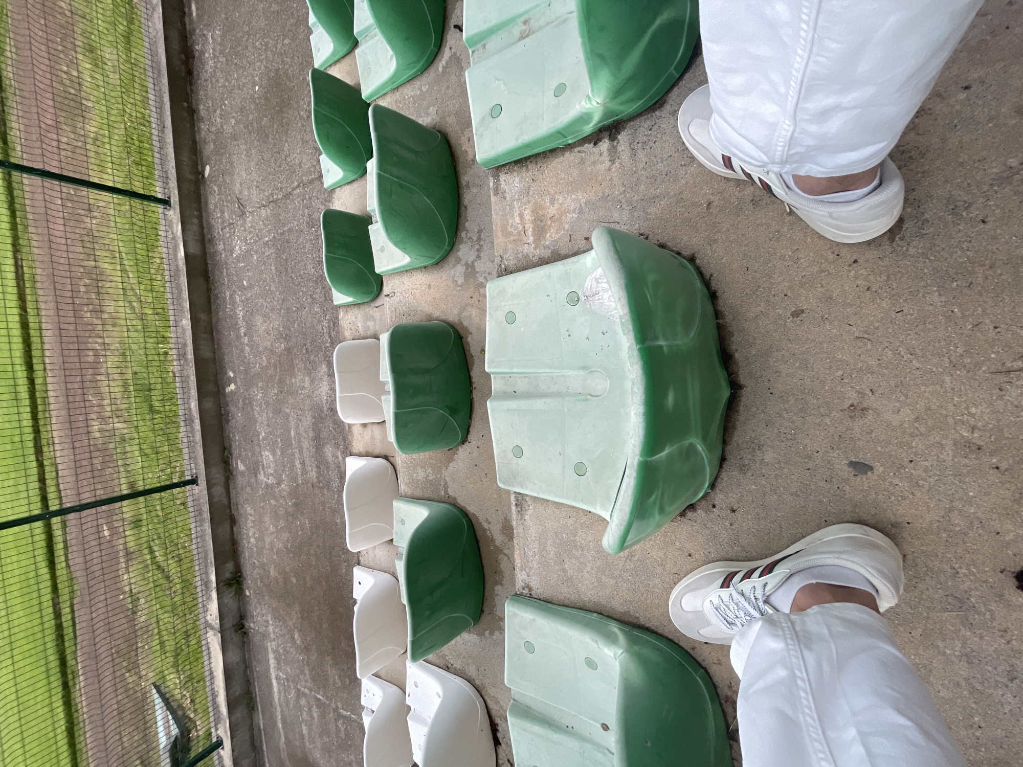 This image, taken in Ihtiman, Bulgaria, captures a high-angle view looking down at a person's feet and the immediate surroundings of a sports stadium or outdoor seating area. The primary subjects are the lower legs and feet of a person, clad in white track pants and white sneakers featuring distinctive red and green striped accents on the sides.
The setting consists of rows of empty, individual plastic seats. Most seats are a vibrant green, with some appearing in a lighter, faded green or white, and they are mounted on a worn concrete floor. To the left, a dark green wire mesh fence separates the spectator area from a grassy field or track, which is visible in the background. The fence has a darker horizontal bar running across it.
The activity depicted is simply the presence of a person standing or walking within the stands. The time of day appears to be daytime, indicated by the natural light, which is soft and diffused, suggesting an overcast sky or a late afternoon. There are no notable shadows or specific weather conditions evident. The overall impression is of a functional, possibly aged, outdoor sports facility. No visible text is present in the image.
