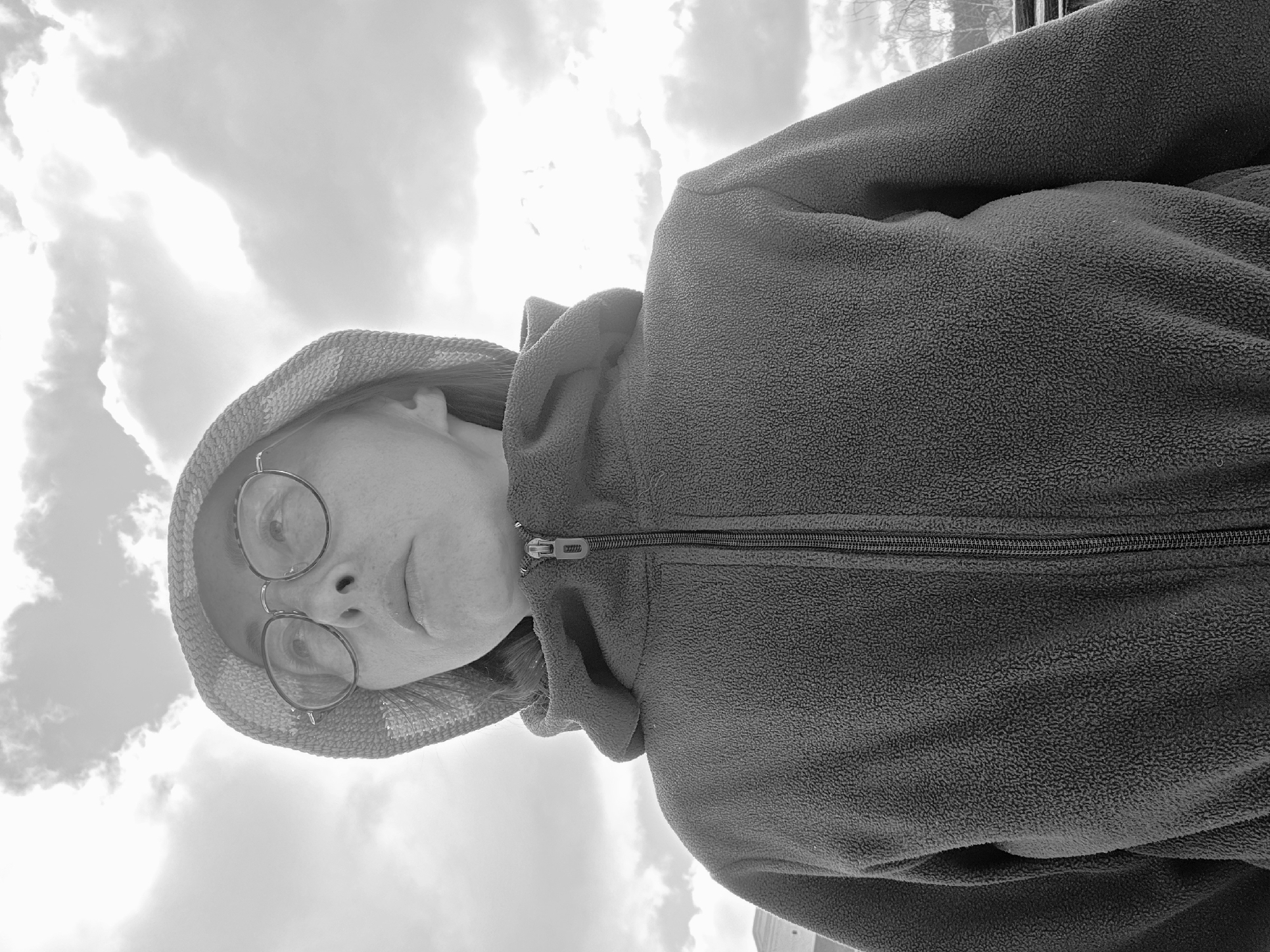 This black and white image is a rotated portrait of a person, viewed from a low angle against a bright, cloudy sky. The subject, who appears to be female, looks directly towards the viewer with a neutral expression. She wears round-rimmed glasses and a textured hat or hood. Her attire includes a dark, fleecy hooded jacket with a visible zipper running down the front. The texture of the fleece is clearly discernible.
The background is dominated by a bright sky filled with scattered clouds, with some areas exhibiting intense light, suggesting strong daylight. The composition emphasizes the person's face and upper body against the expansive sky. There are no specific landmarks or discernible text in the image, though the context indicates the location is Chaikovsky, Russia. The image is oriented with the subject's head to the left and feet to the right, as if the photo was taken with the camera held sideways and pointed upwards.