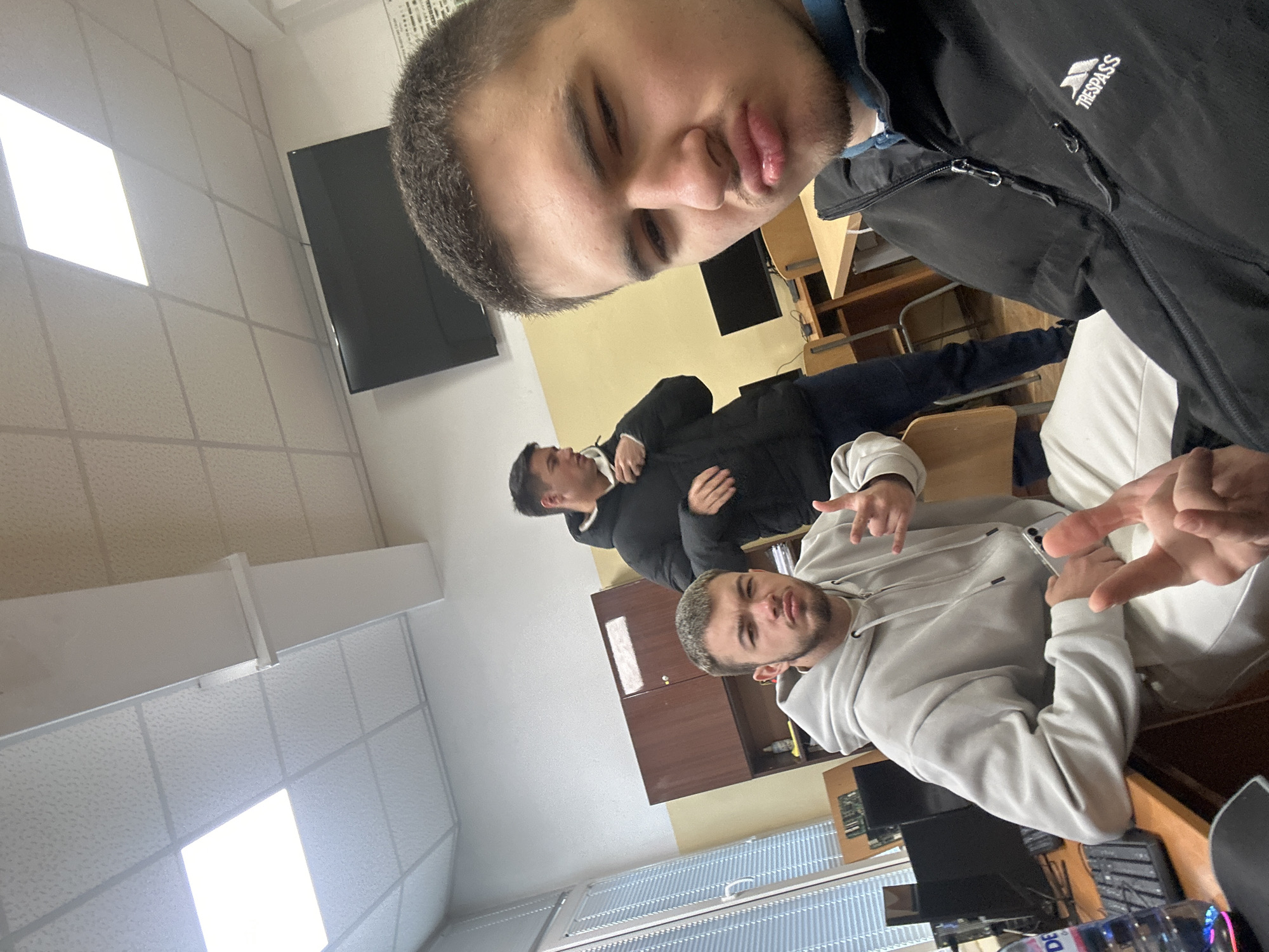 Three men are in an indoor setting, possibly a classroom or office. The man closest to the camera is wearing a black jacket with a "TRESPASS" logo. He is making a pouting face. Another man is seated in the middle wearing a light gray hoodie. He is making a hand gesture. The third man in the background is wearing a black jacket. The room contains desks, chairs, computers, and a cabinet. The ceiling has fluorescent lighting panels. There is a window with blinds in the background. The location is unconfirmed to be Sofia, Bulgaria.