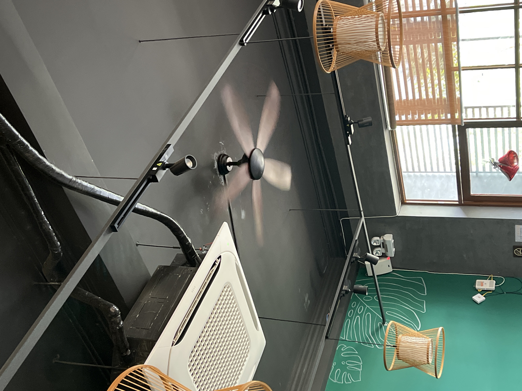 The image shows an interior view in Thuận An, Vietnam. A dark gray ceiling houses a rotating ceiling fan, mounted flush to the ceiling. Track lighting with black spotlights runs along the ceiling, complemented by two hanging lampshades made of light-colored wood or wicker. An air conditioning unit, primarily white, is installed on the ceiling, with associated ductwork visible. A window with wooden blinds is partially visible, and a green wall with white tropical leaf motifs is also present. The scene is well-lit, with natural light coming through the window.