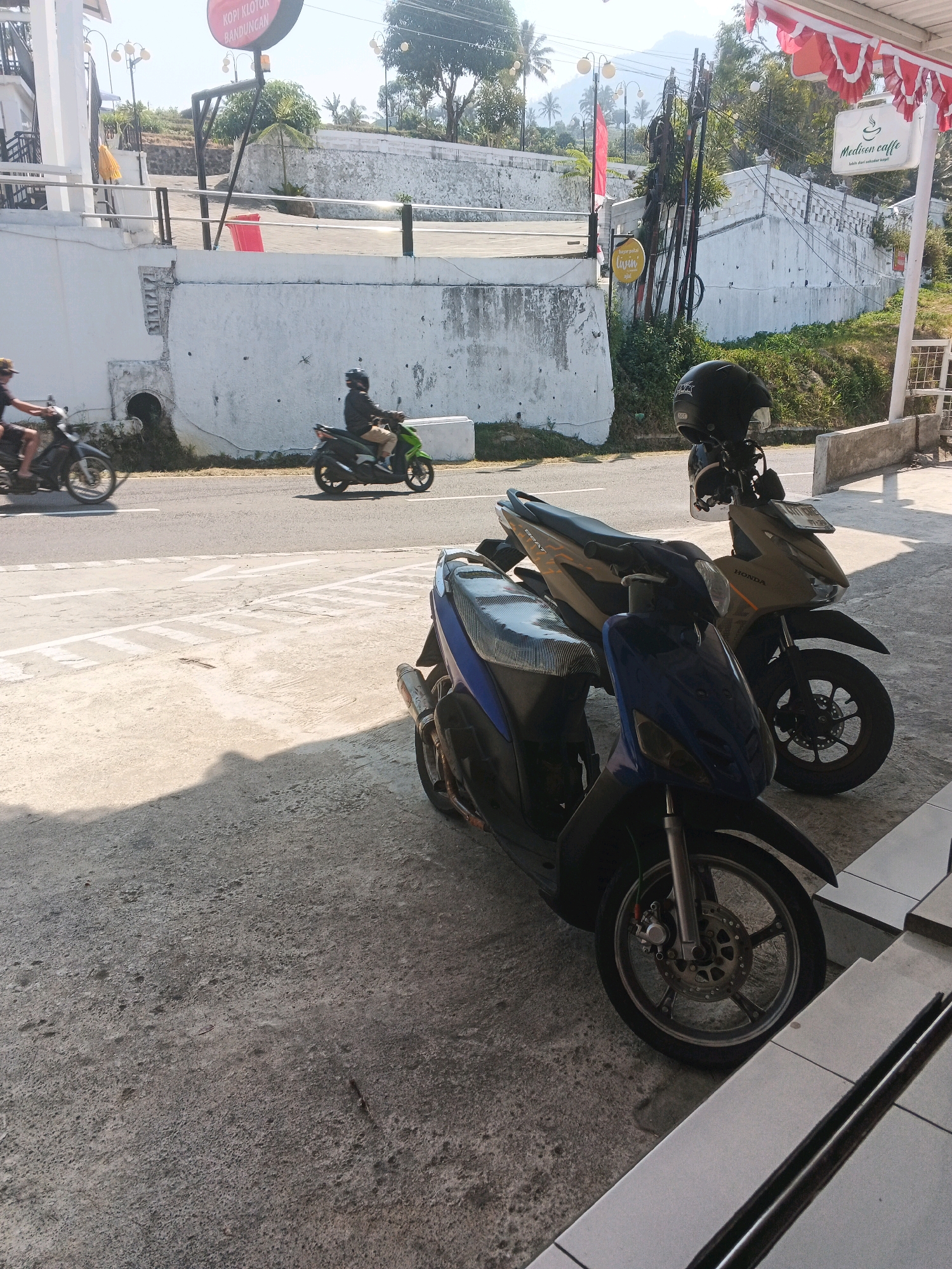 This image captures a street scene in Pakopen, Indonesia, under bright daylight conditions. Two scooters are parked on a concrete surface in the foreground. The scooter on the left is predominantly blue with a black and patterned seat. The scooter on the right is a matte khaki color, with "HONDA" branding visible on its side panel and a black helmet resting on its handlebars. In the background, a road is visible with two people riding scooters. One scooter is black and being ridden by a person wearing a dark shirt and light-colored pants, moving from left to right. Another scooter, colored green, is also moving from left to right, ridden by a person in a dark jacket and light pants. A white concrete wall with some damage and signs is present behind the road. Further in the background, there are trees and a hillside, with some buildings and infrastructure visible, including what appears to be a red sign with white text at the top left. Red and white striped bunting is draped over a structure on the right side of the frame, near a sign that reads "Medusa Caffe". The overall environment suggests a commercial or roadside area.