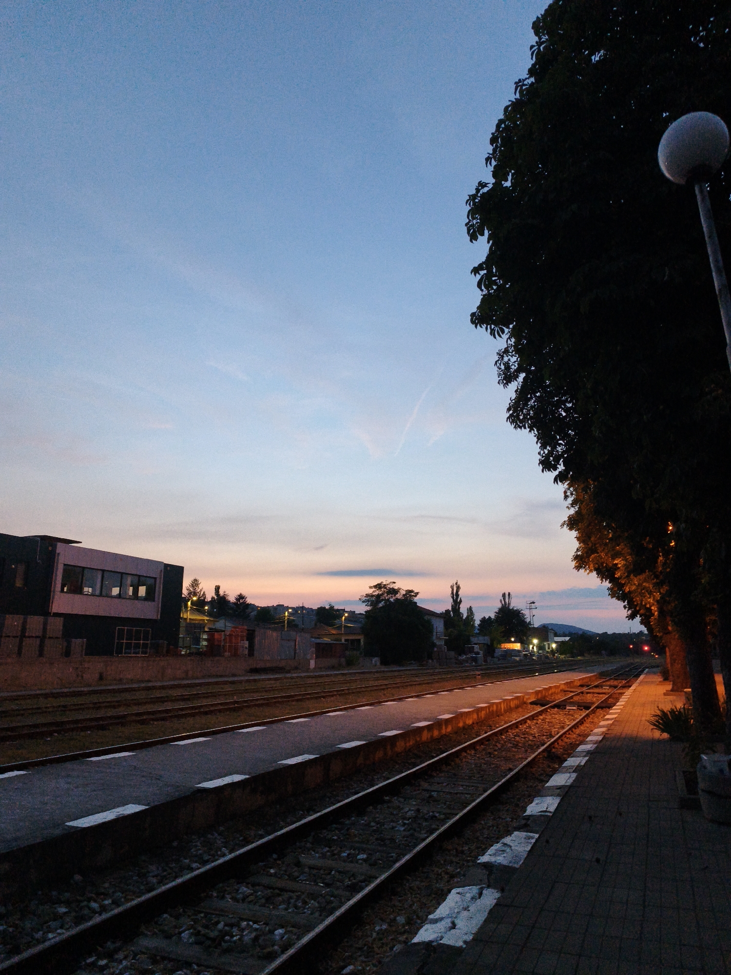 The image depicts a train station at dusk in Lovech, Bulgaria. The sky is a gradient of pale blue and pastel pinks, with faint contrails visible. On the left, a modern building with large windows is partially visible, along with some construction materials. The foreground is dominated by train tracks and a platform. The tracks run diagonally into the distance. To the right, a row of trees casts shadows, with the leaves illuminated by warm light, possibly from street lamps. A spherical street lamp is visible in the upper right corner, and further down the platform, the light from other lamps casts a golden glow on the tracks and pavement. The scene appears deserted, with no people or ongoing activity. The overall impression is one of quiet anticipation at the end of the day.