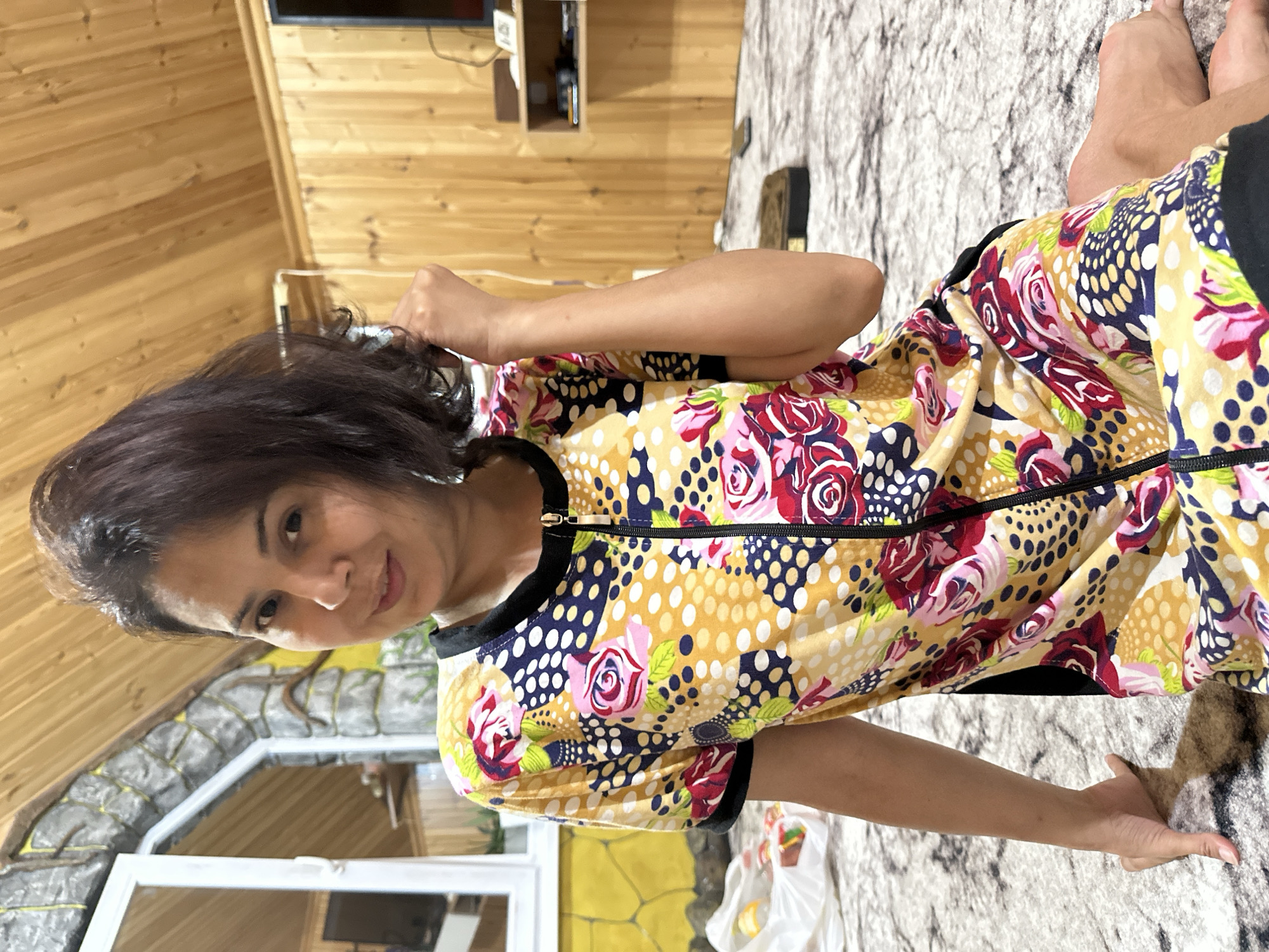 A woman with dark hair is sitting on a patterned carpet in a room with wood-paneled walls and a wooden ceiling. She is wearing a short-sleeved zippered dress with a colorful floral and polka dot pattern. She is looking towards the camera with a slight smile and has her right hand raised, touching her hair. To the left of the woman, a window is visible with a white frame, and beyond it, a textured wall with stone-like accents. A white plastic bag with some items inside is placed on the carpet near the window. In the background, there is a shelf unit with some objects on it. The environment appears to be indoors, possibly a residential space.