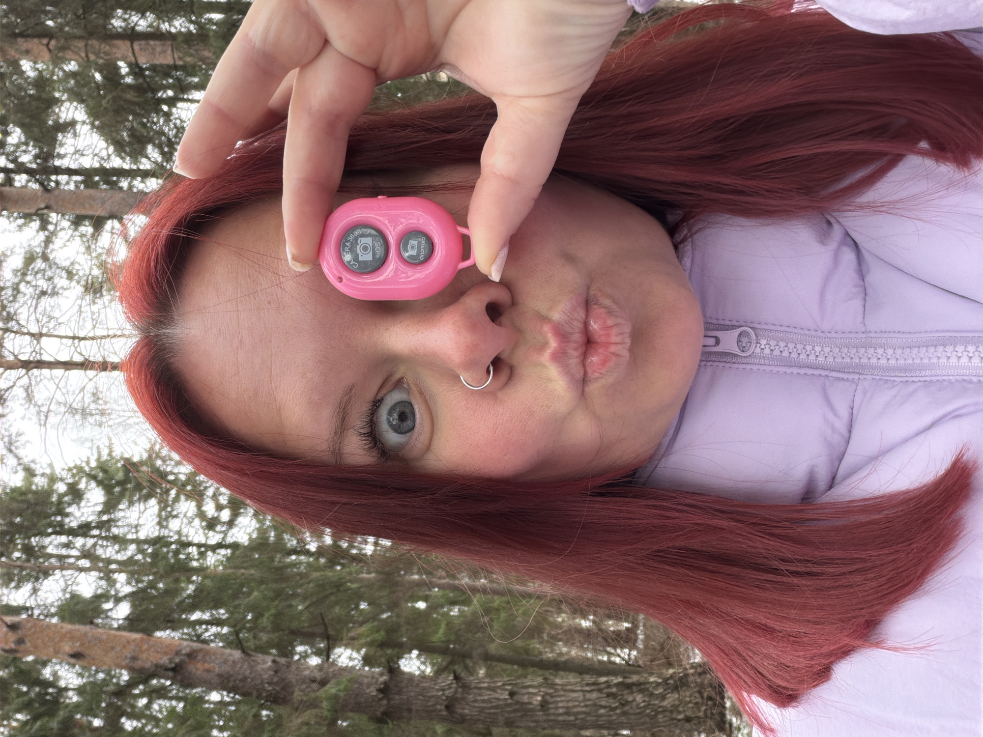 A woman with auburn hair, a nose ring, and light blue eyes is holding a pink, two-button device in front of one eye. The device is a Bluetooth remote for a camera, labeled with camera symbols and the words "CAMERA 360" and "ANDROID IOS" around the buttons. The woman is pouting, and she's wearing a lavender-colored puffer jacket. The background is a blurry mix of trees, suggesting an outdoor setting.
There is no information in the picture to indicate that the location is in the city of Chaikovsky, Russia.