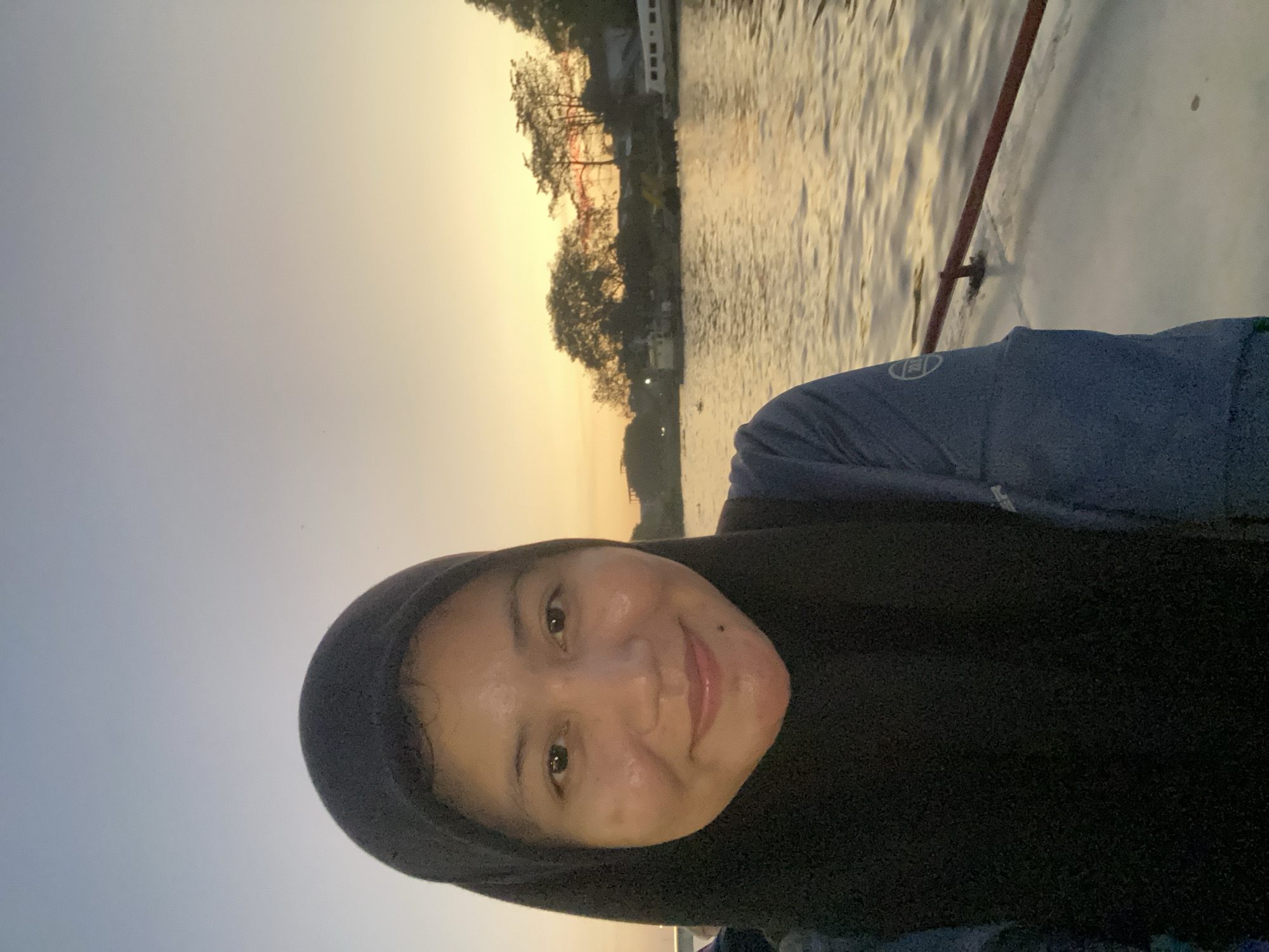In Banjarmasin, Indonesia, a woman wearing a black hijab and a blue shirt is taking a selfie on a boat. She is smiling at the camera, with the sun setting behind her, casting a warm glow on the water. The tranquil scene is enhanced by the calm waters of the river, reflecting the golden hues of the sunset. The background shows lush green trees and buildings along the riverbank, giving a glimpse of the city's charm. The boat she is on appears to be a local mode of transport, commonly used to navigate the numerous rivers and waterways of Banjarmasin.