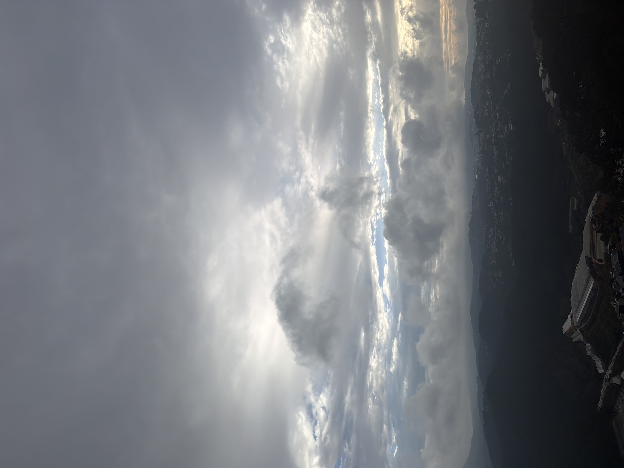 This is a high-angle, landscape photograph taken from a vantage point above the city of Xuân Thành, Vietnam, likely during an overcast or dawn/dusk period. The sky dominates the upper two-thirds of the image, filled with a complex layering of clouds. Heavy, dark gray clouds occupy the upper portion, with lighter, broken clouds and patches of sunlight breaking through lower down, particularly towards the right side where a hint of warm sunset or sunrise colors is visible.
Below the sky, a vast expanse of rolling hills and valleys stretches out, blanketed in shadow. This is the cityscape of Xuân Thành, appearing as a dense collection of buildings and structures nestled amongst the darkened landscape. The town's lights are not clearly discernible due to the low light conditions, but the pattern of habitation is evident. In the foreground and lower right quadrant, several large, modern structures, appearing to be greenhouses with white sheeting, are visible on the slopes, indicating agricultural or horticultural activity. The overall impression is one of a sprawling, somewhat subdued urban environment set against a dramatic, cloud-filled sky. No people are visible in the image.