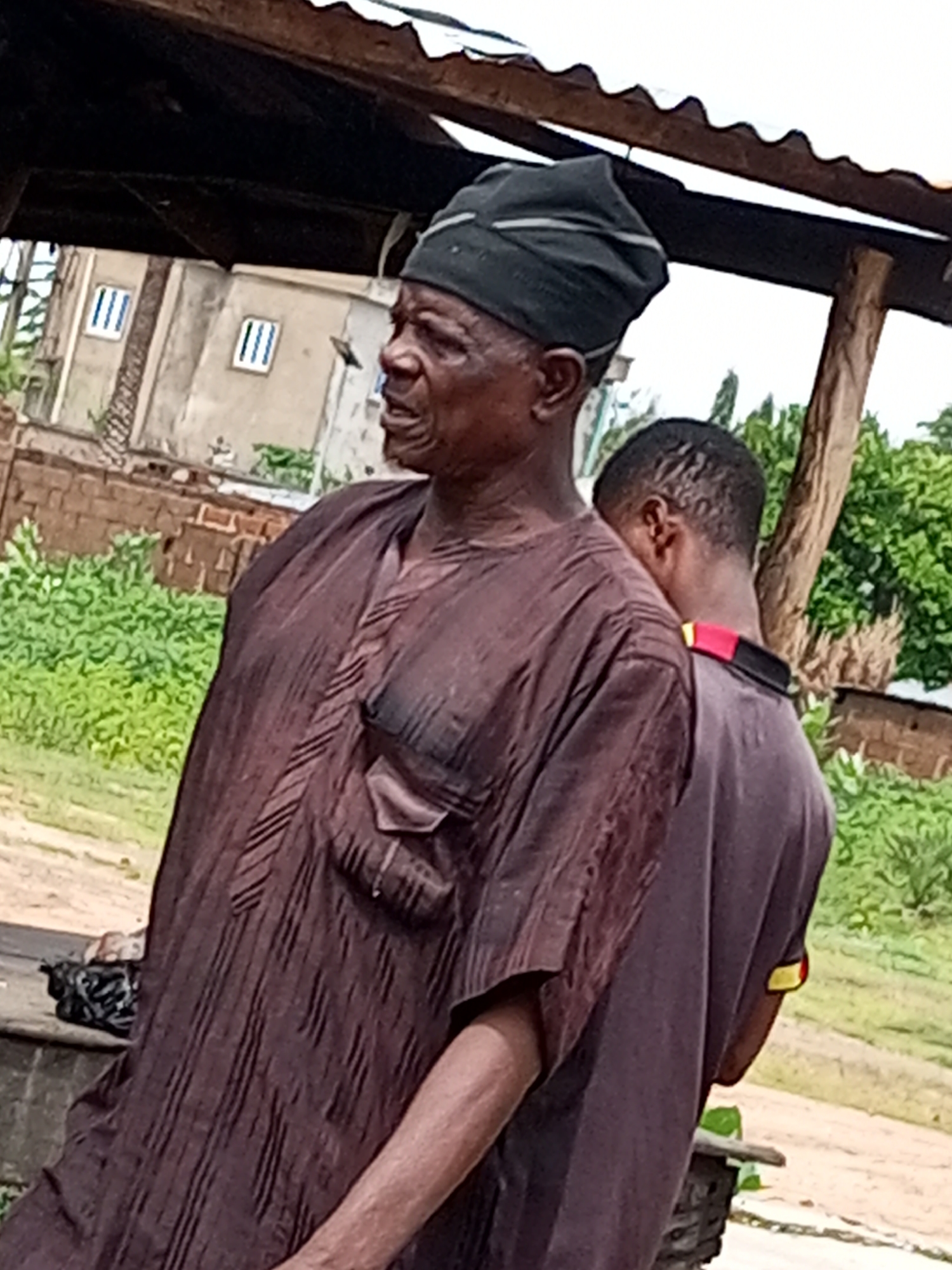 Two individuals are visible outdoors in Garko, Nigeria. The primary subject is an elderly man in the foreground, facing to the left of the frame. He is wearing a dark, possibly black, headwrap and a long, dark brown garment with a textured pattern. The garment appears to have a patch pocket on the left side. His facial features are visible, and he is looking away. Behind him and slightly to his right is a younger individual, facing away from the camera and towards the right. This person is wearing a dark t-shirt with a red and yellow stripe on the collar. The background shows a partially constructed building with multiple windows, brickwork, and dense green foliage. A corrugated metal roof supported by wooden beams is visible above the individuals, suggesting a covered outdoor area. The ground appears to be dirt or gravel. A black plastic bag is present on a ledge to the left of the foreground subject. The overall impression is a candid outdoor scene in a semi-urban or rural environment.