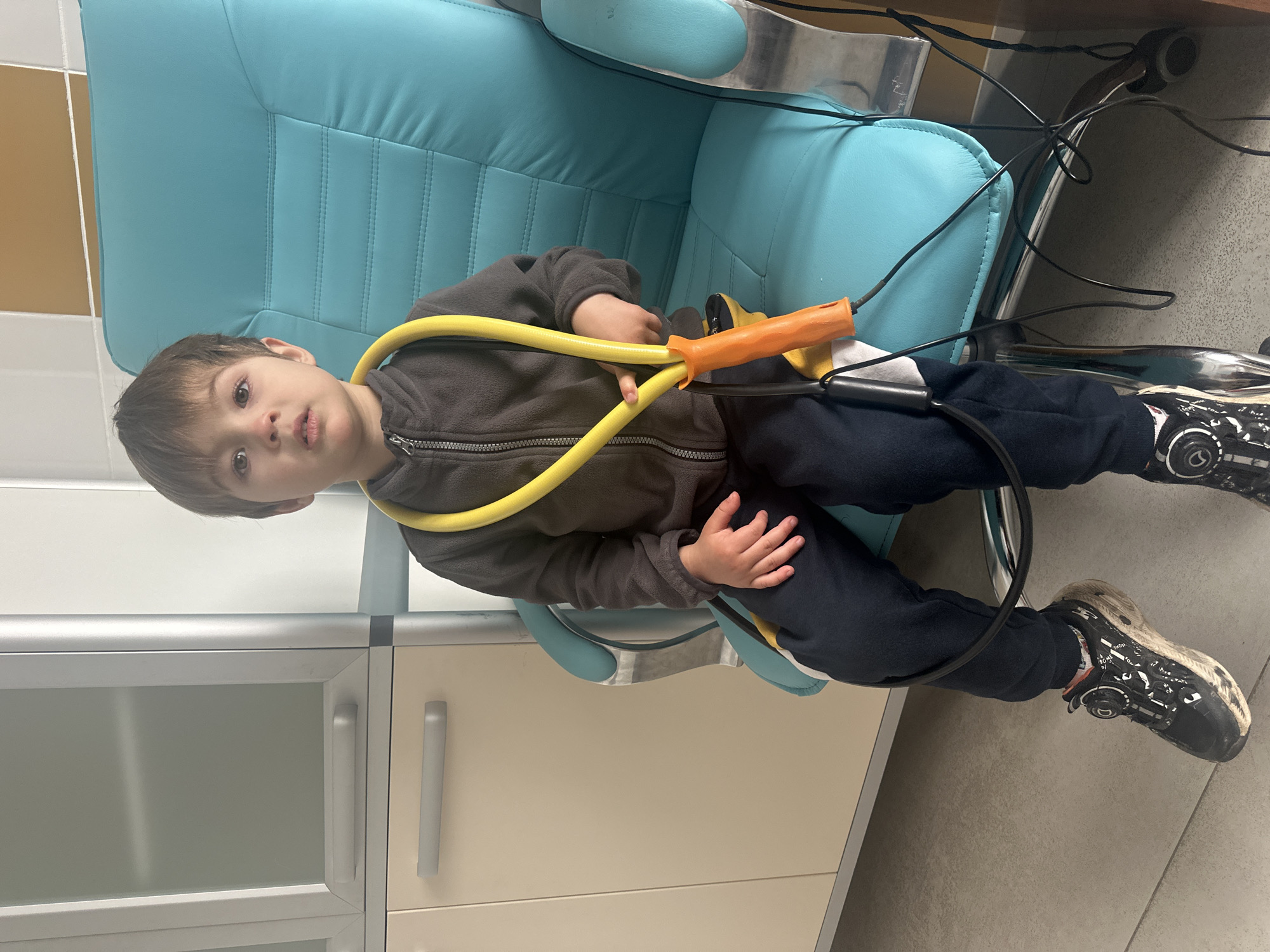 A young boy is captured sitting on a modern turquoise office-style chair, looking directly at the camera with a calm expression. He appears to be a toddler or preschooler, dressed in a dark grey zippered fleece jacket and dark blue sweatpants. He holds a bright yellow, flexible tube-like object with an orange handle, possibly a toy or exercise equipment, across his lap with both hands.
The setting appears to be an indoor room, likely a professional or institutional environment such as a clinic, waiting room, or office, rather than a home. This is suggested by the design of the chair and the backdrop of light beige and light grey cabinets or drawers with sleek gray handles. Black cables or wires are visible behind and to the side of the chair, some coiled on the floor or dangling, hinting at electronic equipment nearby. The floor is a light gray tile. The boy is wearing black and white sneakers, which show some signs of wear. The image is taken from a slightly elevated perspective, looking down at the boy. No visible text is discernible in the image. The location is Khimki, Russia.