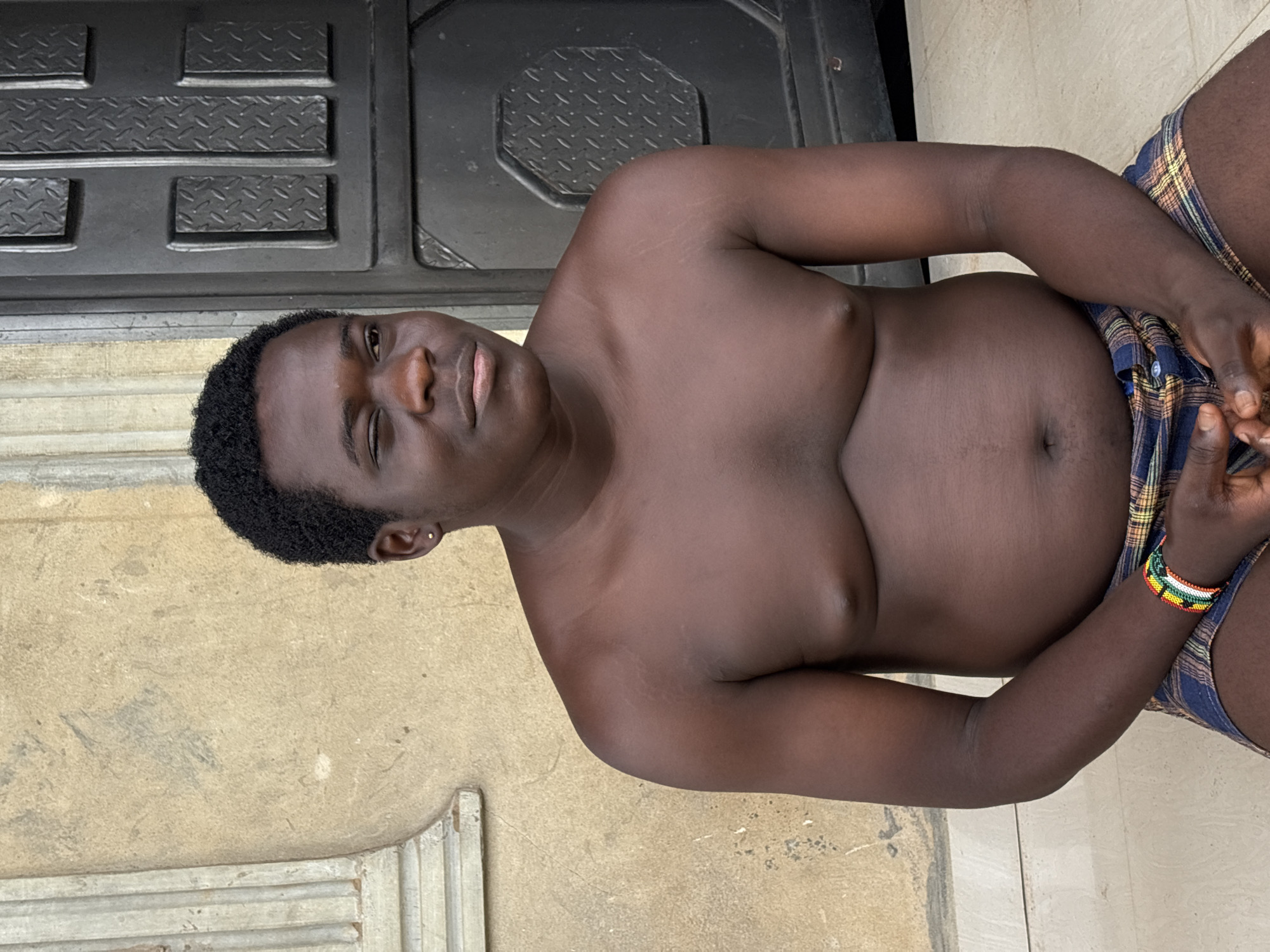 The original image, presented rotated 90 degrees counter-clockwise, depicts a young, dark-skinned person, likely male, reclining outdoors. In its intended vertical orientation, the person is shown from the waist up, looking directly at the viewer with a relaxed expression and a slight smirk. He has short, dark, curly hair and is shirtless, revealing his bare torso and midsection. He wears plaid shorts in shades of blue, yellow, and brown, with his hands clasped together and resting on his left leg.
Notable details include a small, silver-colored stud in his left earlobe. Faint stretch marks are visible on his shoulders and chest. A colorful beaded bracelet, featuring bands of red, yellow, green, white, and black, is worn on his right wrist.
The background consists of a light, textured wall, possibly stucco or concrete, to the viewer's left. Behind the person and to the viewer's right is a dark, possibly metallic or heavy plastic panel with a raised diamond plate pattern, suggesting a utilitarian structure like a door or gate. The ground beneath him appears to be made of light-colored tiles.
The scene takes place during the daytime, indicated by the natural lighting. There is no visible text in the image. The location context for this image is Ikot Ekpene, Nigeria.