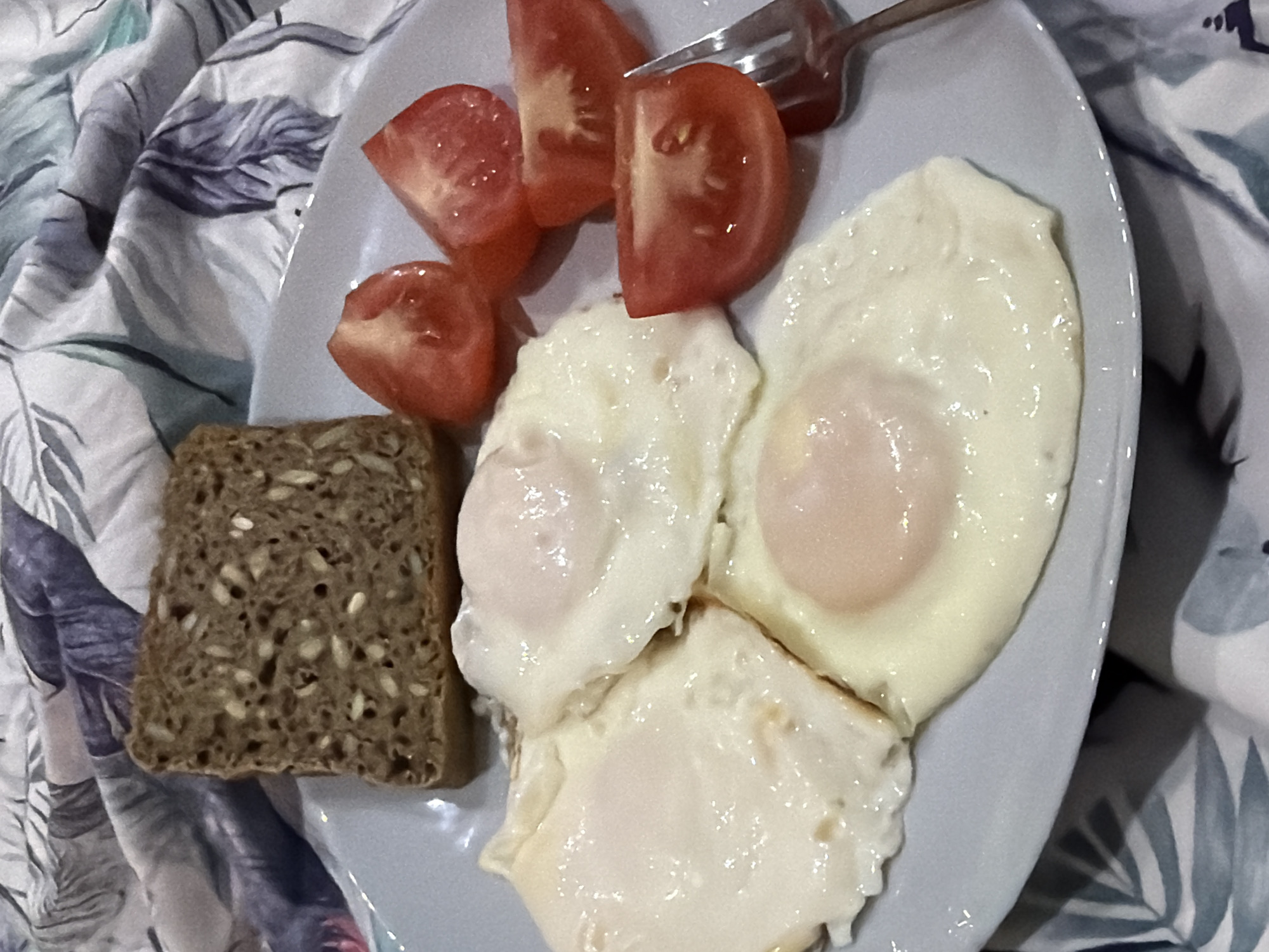 A plate of food sits atop a patterned fabric. The food consists of three fried eggs, several tomato slices, and a piece of dark bread speckled with seeds. A fork rests partially on top of the tomato slices. The plate is light blue. The fabric features a print of stylized leaves in muted blue, gray, and white tones. The location is unknown, and there is no information in the image or prompt to indicate this image was taken in the city of Chaikovsky, Russia. No people are visible.
