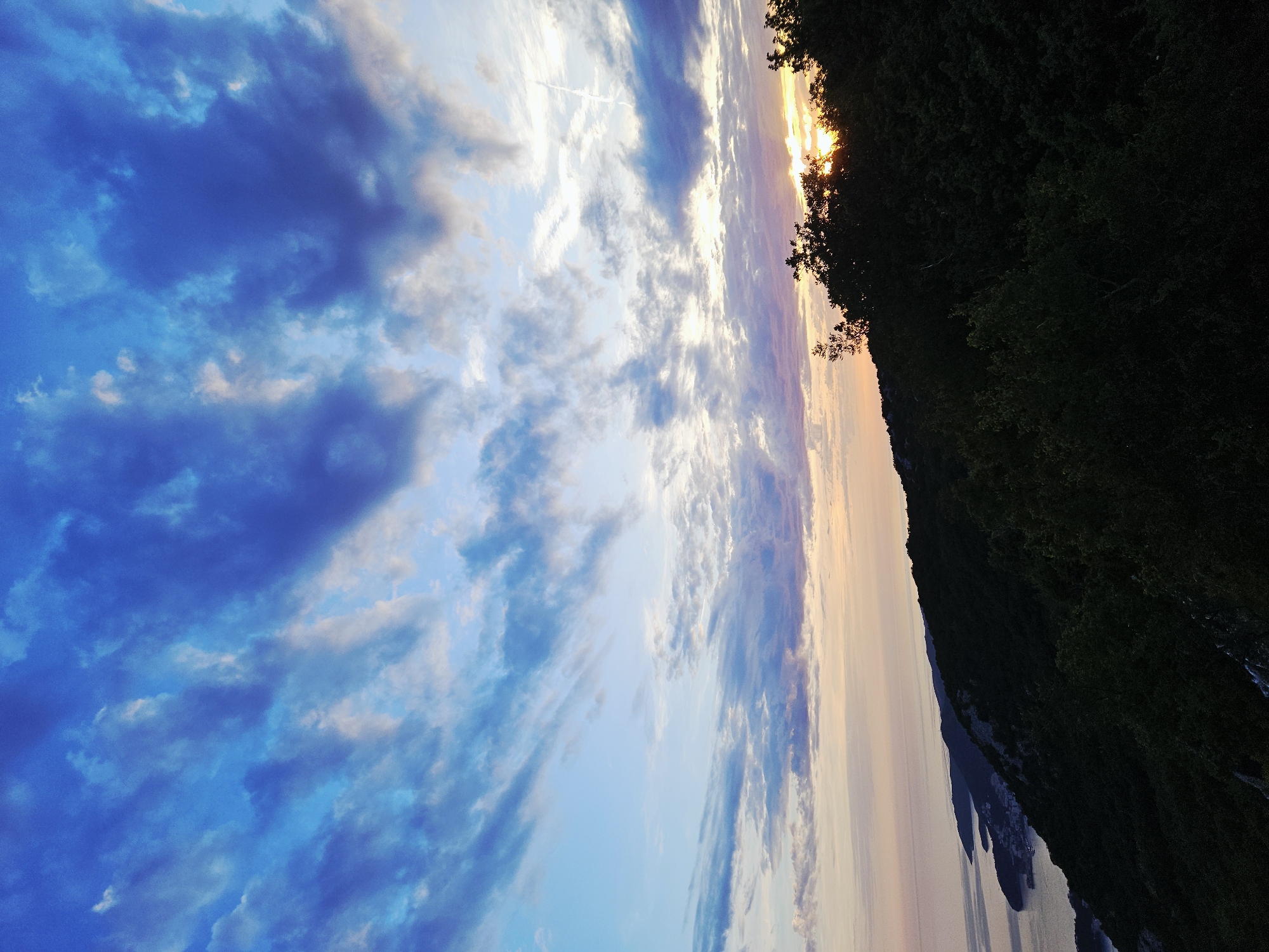 The image captures a coastal landscape at either sunrise or sunset, located in the city of Unknown, Unknown. The upper portion of the sky is a deep blue, heavily populated with layered clouds ranging from dark blue-grey to bright white. Towards the horizon, the sky transitions into lighter blues, pinks, and oranges, illuminated by a low-lying sun. A thin, white contrail is visible in the upper right quadrant of the sky.
The sun itself is a bright orange glow, partially obscured by the dark silhouette of a densely forested hill on the right side of the frame. Below the sky, a calm body of water, likely the sea, extends across the mid-ground on the left. A rugged coastline with multiple hills and bays runs along the left side, separating the water from the foreground landmass. A small town or settlement with visible structures is nestled within a bay along this coastline in the mid-distance. The immediate foreground consists of a dark, tree-covered hillside, primarily composed of dense foliage and dark rock formations, which rises diagonally from the bottom of the image.