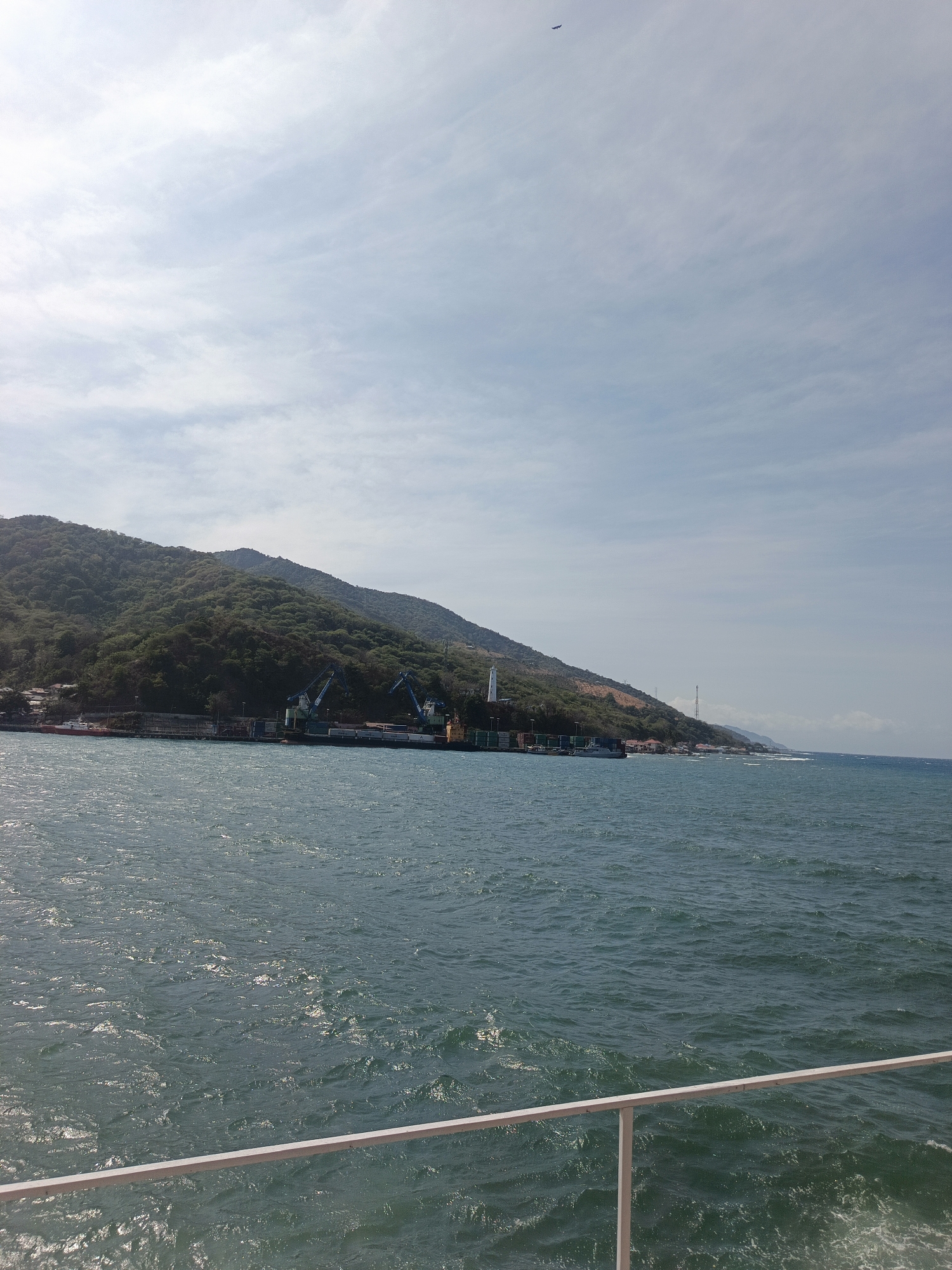 A horizontal, medium shot captures a view from a boat looking towards a coastal area on a sunny day. The foreground is dominated by the rippling turquoise water of the sea, with a white railing visible at the bottom edge of the frame, suggesting the viewpoint is from a vessel. In the midground, a shoreline is lined with a port facility. Several large cargo ships are docked, and industrial cranes, two of which are prominent in blue, are positioned along the quay. Shipping containers are stacked in neat rows on the docks. Behind the port, a densely forested, green hillside rises steeply, forming a natural backdrop. The sky above is a pale blue with scattered white clouds, and a faint object, possibly an airplane, is visible in the upper left portion of the sky. The scene depicts a working port situated at the base of a lush, mountainous coastline.