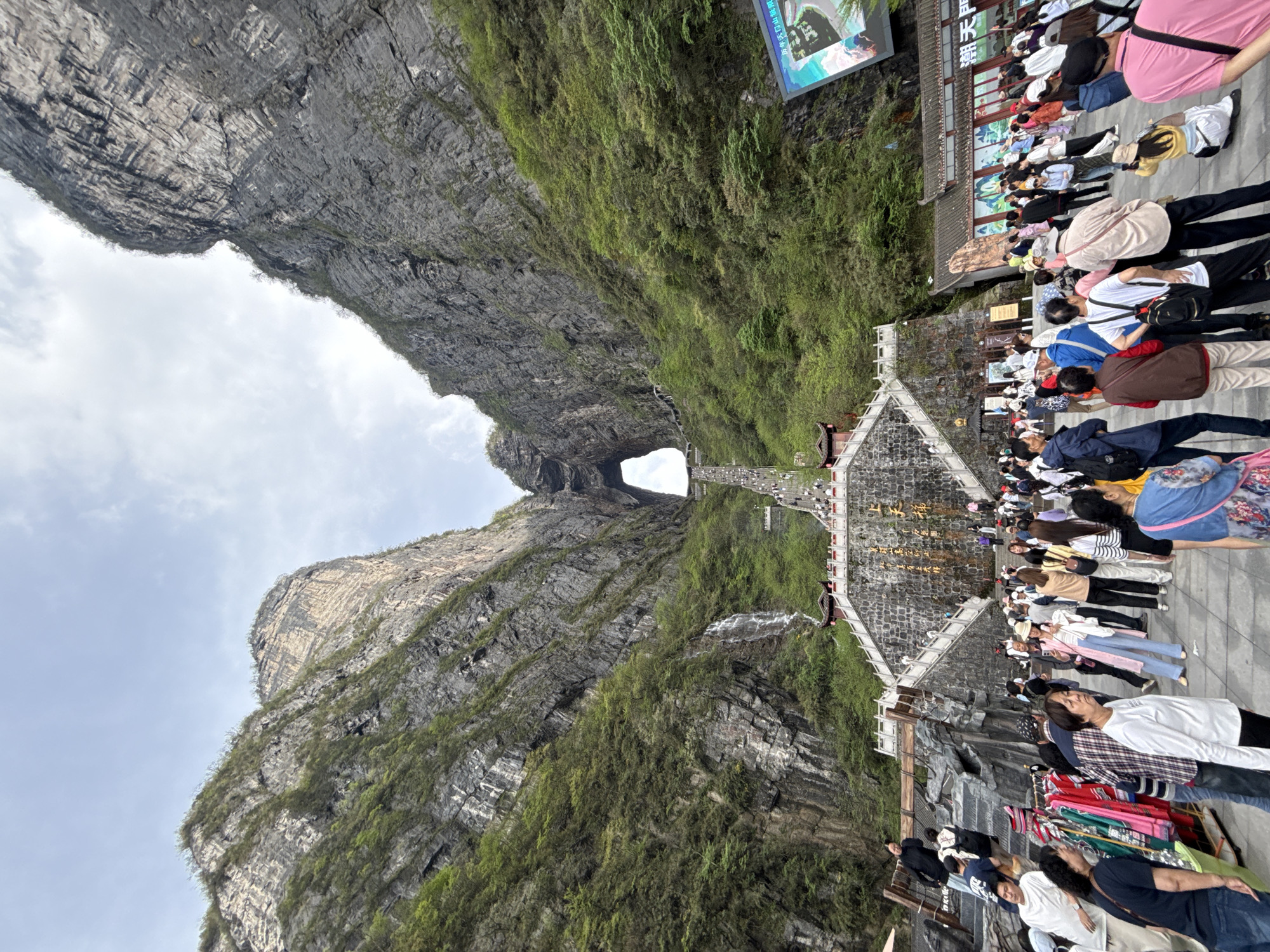 The image shows the Tianmen Cave, or "Heaven's Gate," a large natural arch in Tianmen Mountain, located near Zhangjiajie City, Hunan Province, China. The arch is visible in the distance between the two sides of a large mountain. In the foreground, there is a paved area crowded with tourists. The slope of the mountain is covered in vegetation. The image is taken from below, looking up at the arch and the surrounding area.