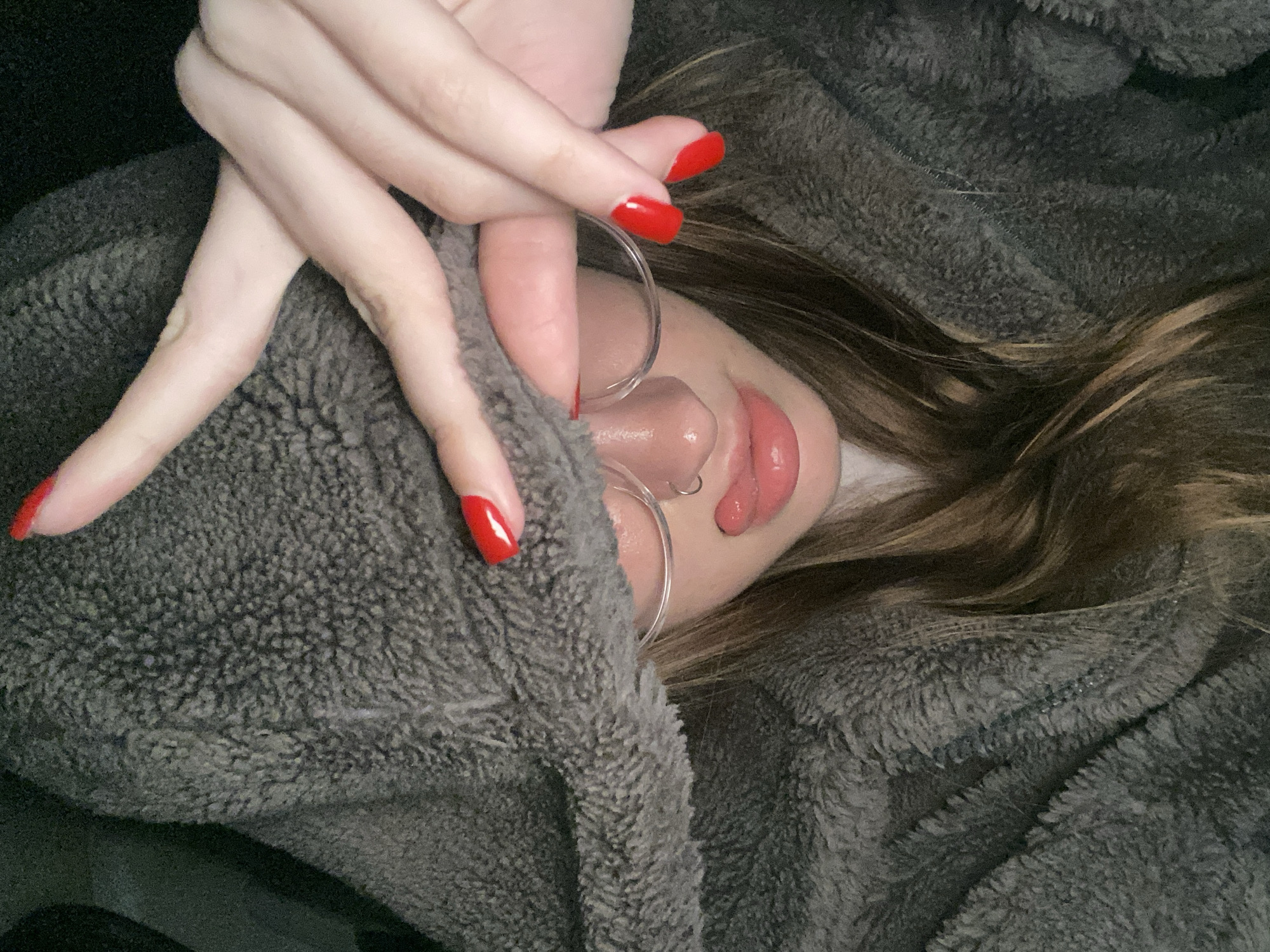 This close-up portrait features a person partially covering their face with their hands. The hands have long fingers with bright, glossy red manicured nails and are intertwined, resting over the upper part of the face, obscuring the eyes and forehead.
Visible facial features include the nose, which has a small hoop piercing on the left nostril, and full, glossy lips that are slightly parted. The person is wearing clear-rimmed glasses that rest on the bridge of their nose. Light brown hair with subtle highlights frames the face, falling onto a soft, plush, grey-colored material, likely a hooded sweatshirt or blanket, which provides a cozy texture and partially envelops the head and neck.
The overall scene conveys a sense of intimacy and comfort, possibly depicting a moment of relaxation, contemplation, or shyness. The lighting is soft, highlighting the textures of the hands, hair, and the fluffy grey fabric. No text is visible in the image.