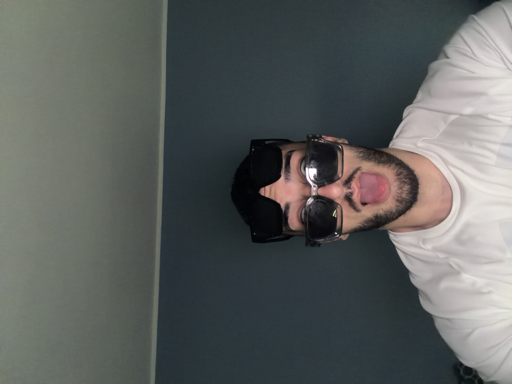 A man with black hair and a beard is wearing sunglasses and sticking his tongue out. He is wearing a white shirt. He is standing in front of a blue wall next to a light grey wall. This photo appears to have been taken indoors and there is no indication of it being taken in Plovdiv, Bulgaria.